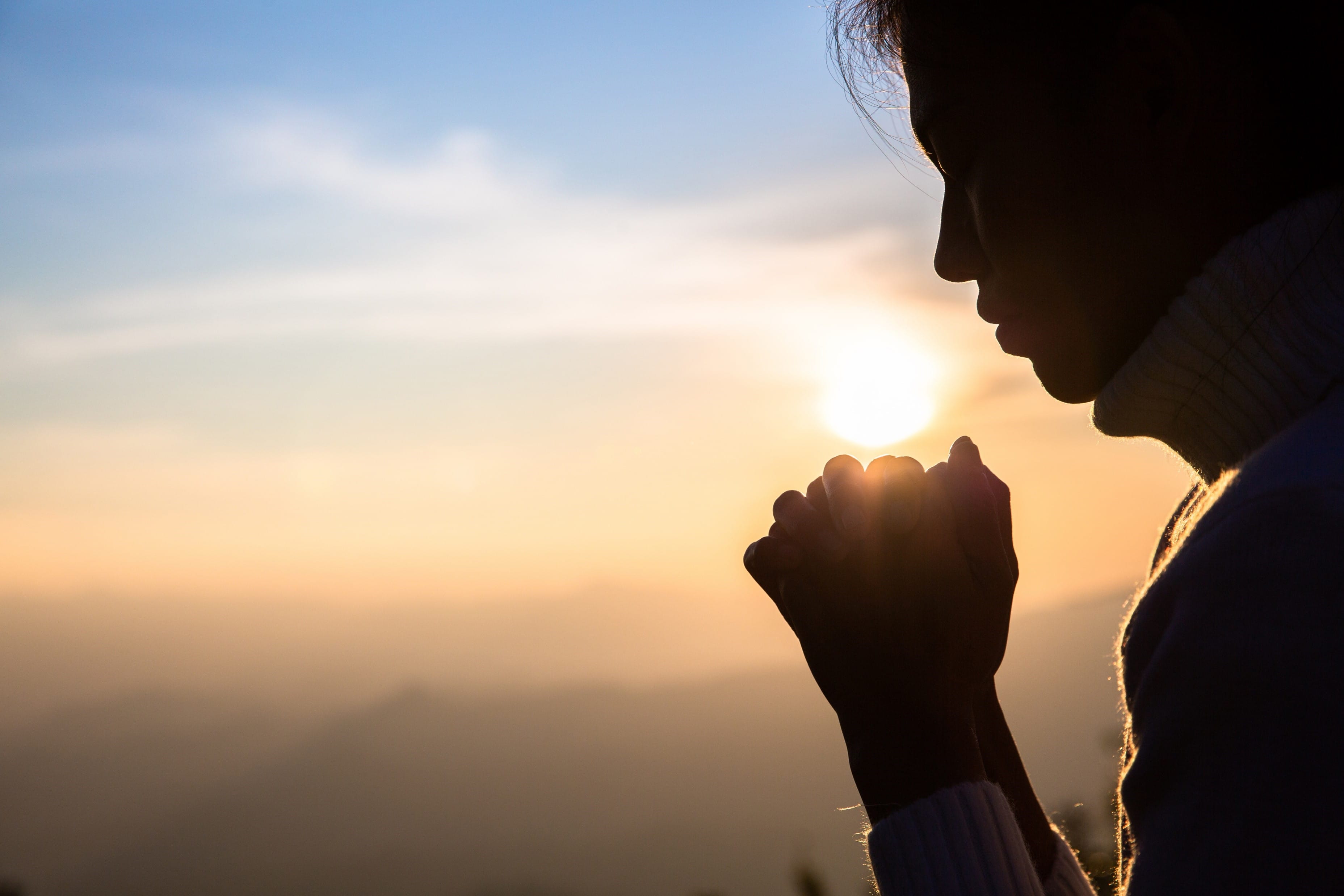 Silhouette of a woman with hands clasped in prayer against a glowing sunrise sky.