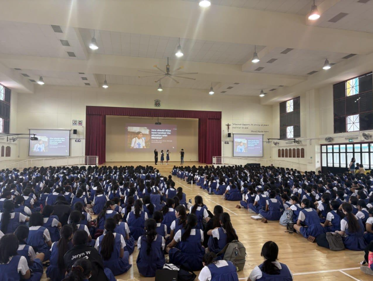 Students in blue uniforms sit on a hall floor facing a stage with presenters and projection screens.