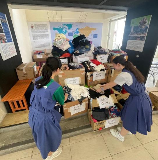 Two girls in school uniforms sort donated clothing into labeled cardboard boxes indoors.