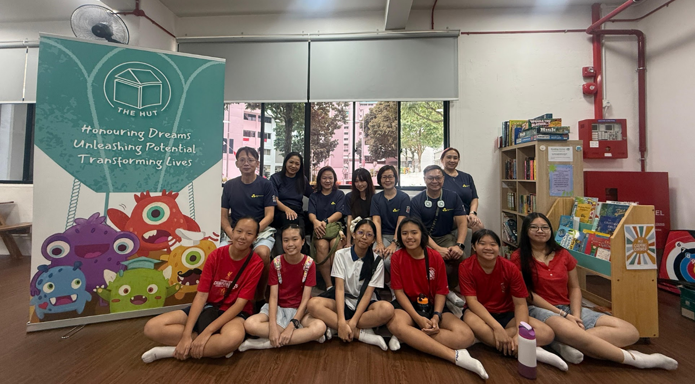 Group of adults and teens posing indoors near a colorful "The Hut" banner and a bookshelf.