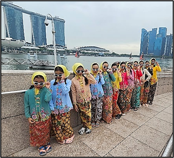 Girls in colorful traditional attire and sunglasses pose in a line by a waterfront with Marina Bay Sands in the background.