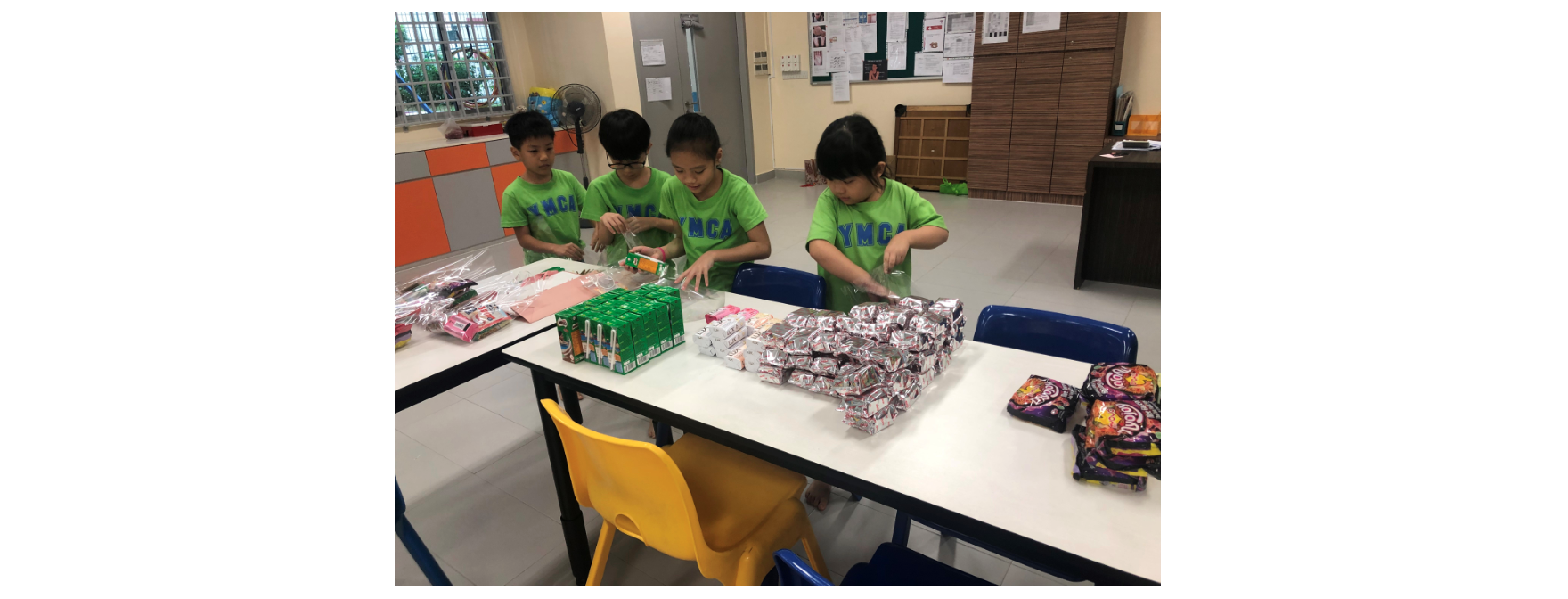 Four children in green YMCA shirts sort snacks and packaged goods on a table in a classroom.