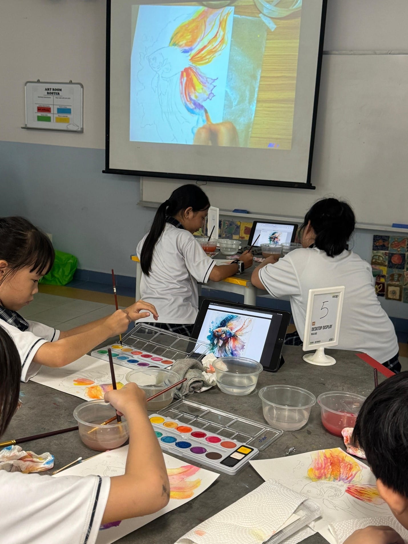 Students in school uniforms paint watercolors at a table, referencing fish artwork displayed on tablets and a projector screen.