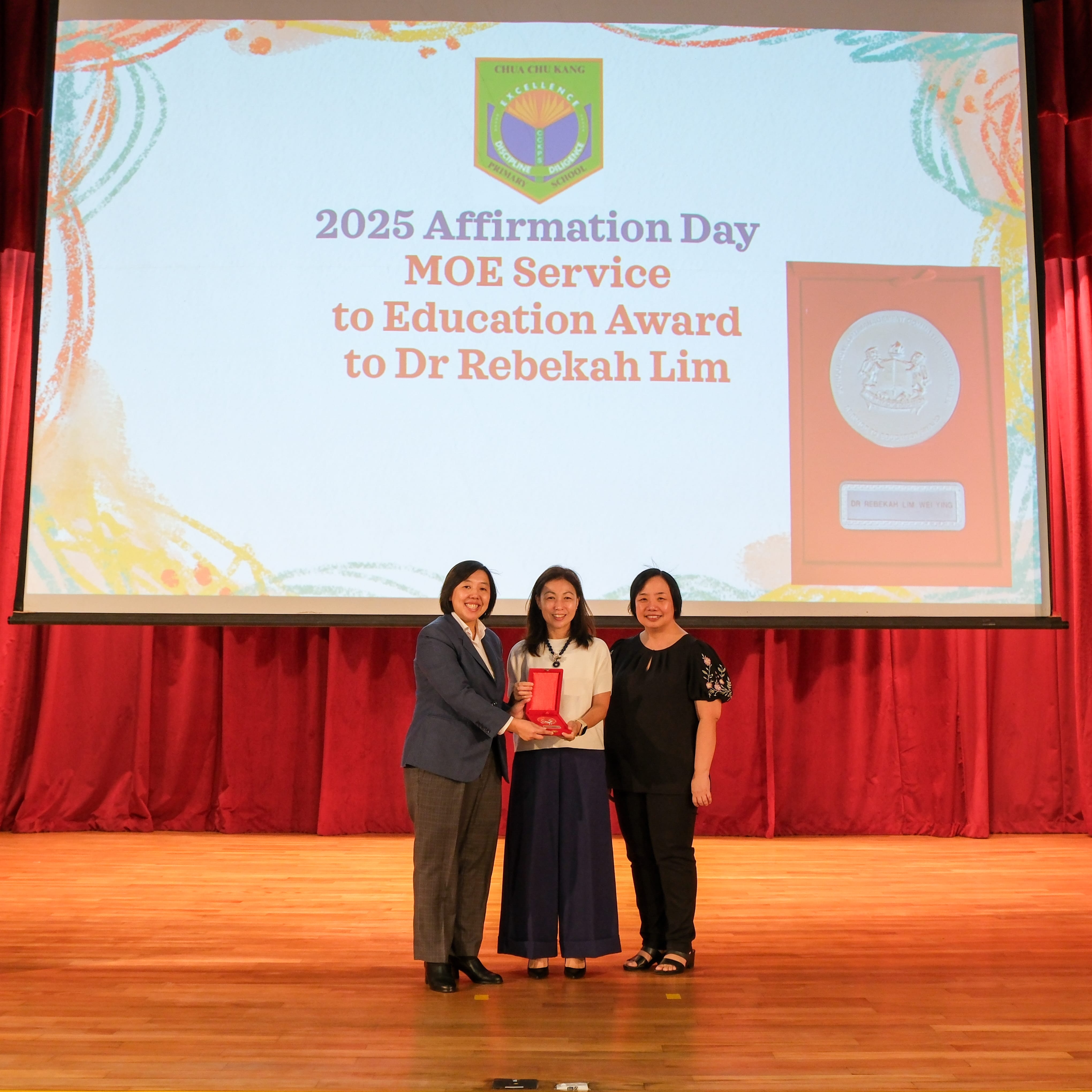 Three women on stage hold a red award case beneath a screen reading 2025 Affirmation Day award to Dr Rebekah Lim