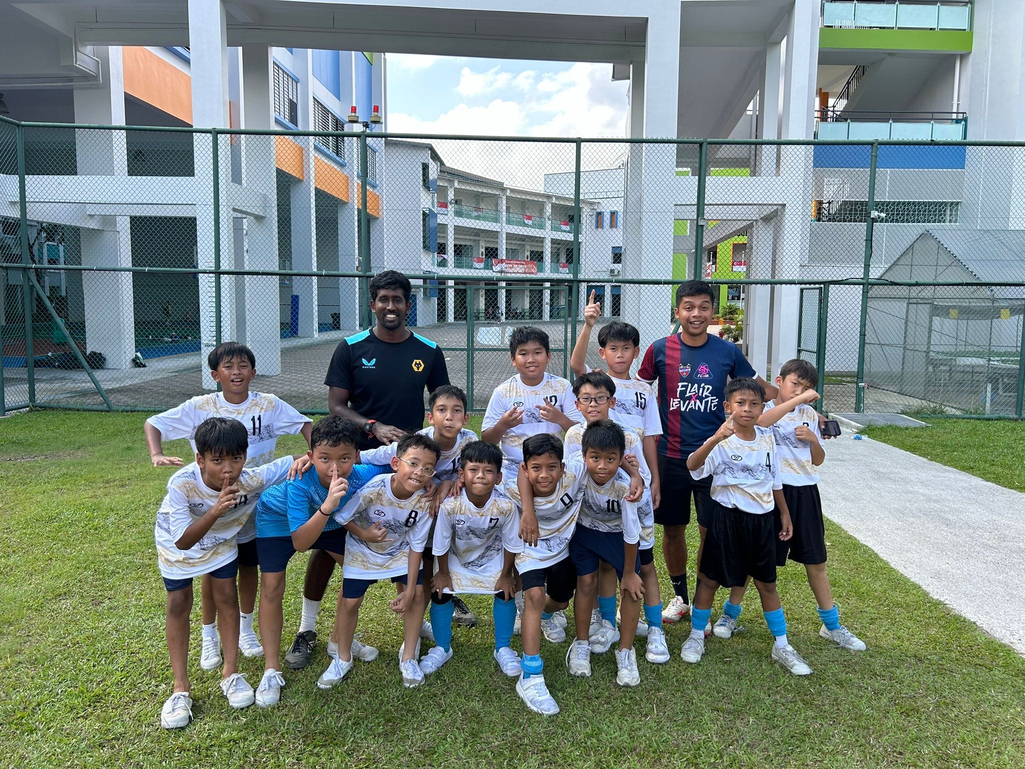 A group of young boys in white soccer jerseys pose with a coach on a grass field outside a school building.