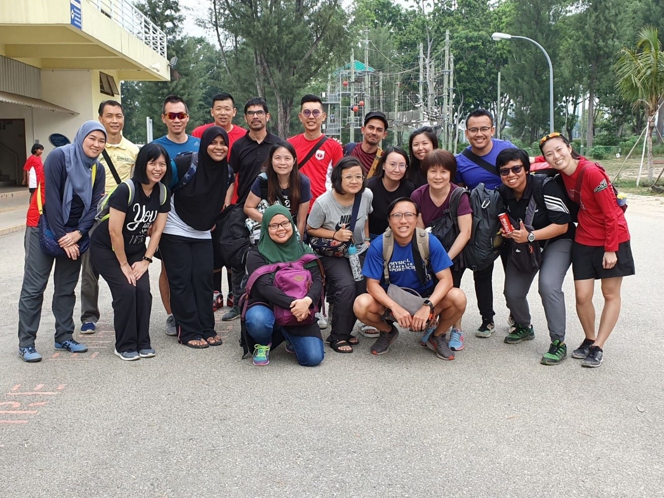 A large group of about 20 people posing together outdoors near trees and a ropes course structure.