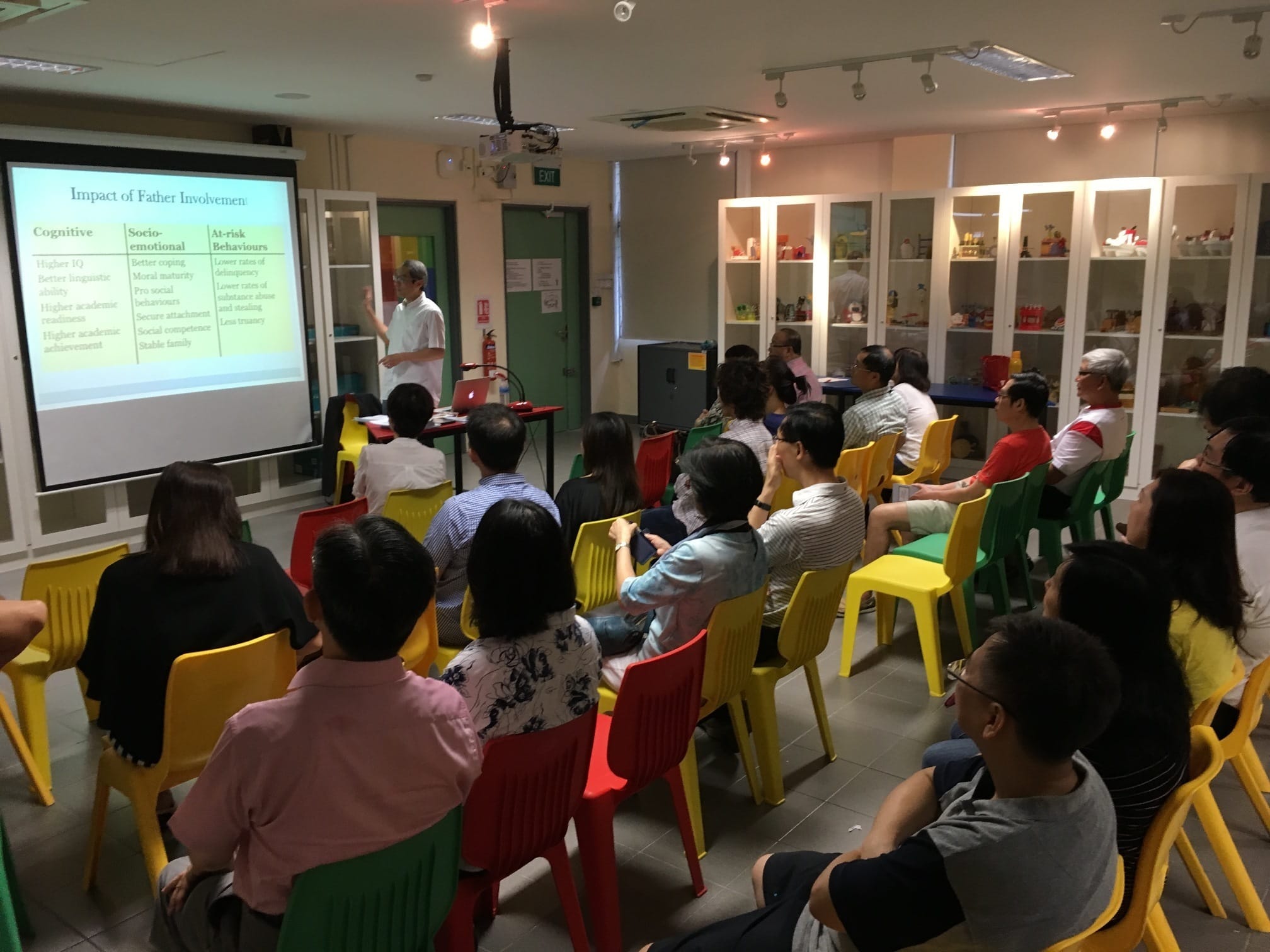 A speaker presents slides titled "Impact of Father Involvement" to a seated audience on colorful plastic chairs.