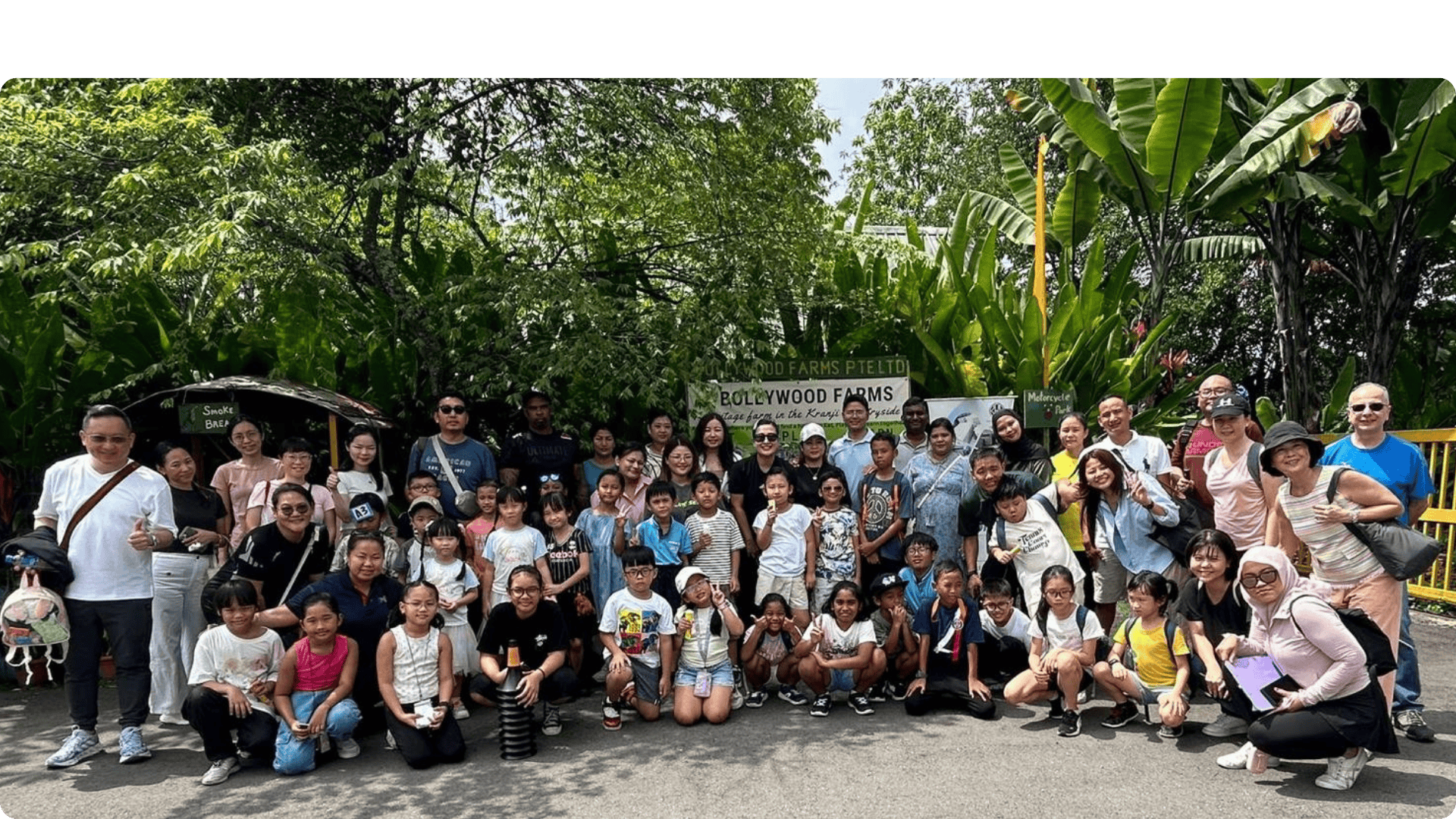 Large group of adults and children posing outdoors in front of a Bollywood Farms sign surrounded by trees.