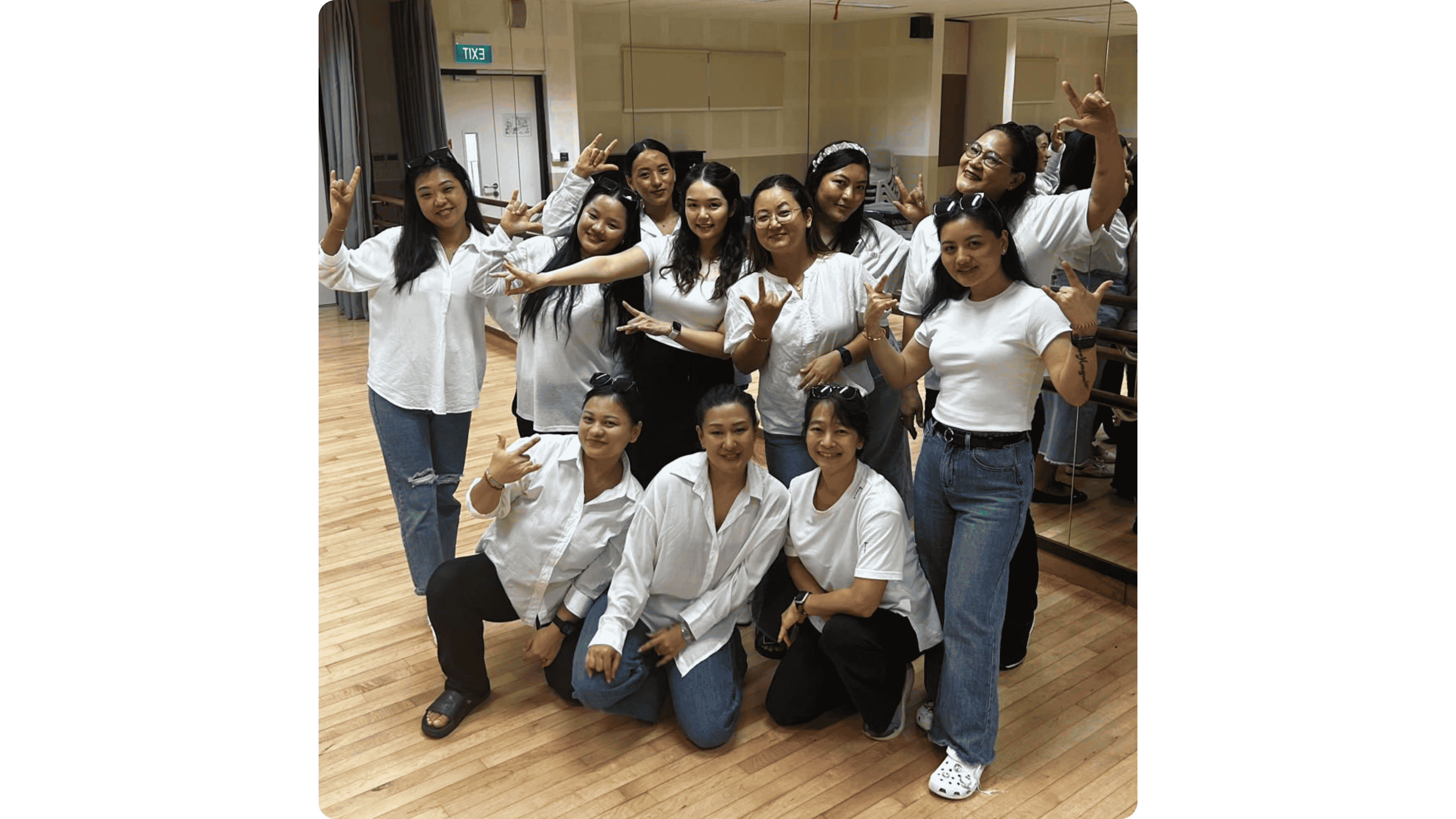 A group of young women in white tops and jeans pose together in a dance studio with wood floors.