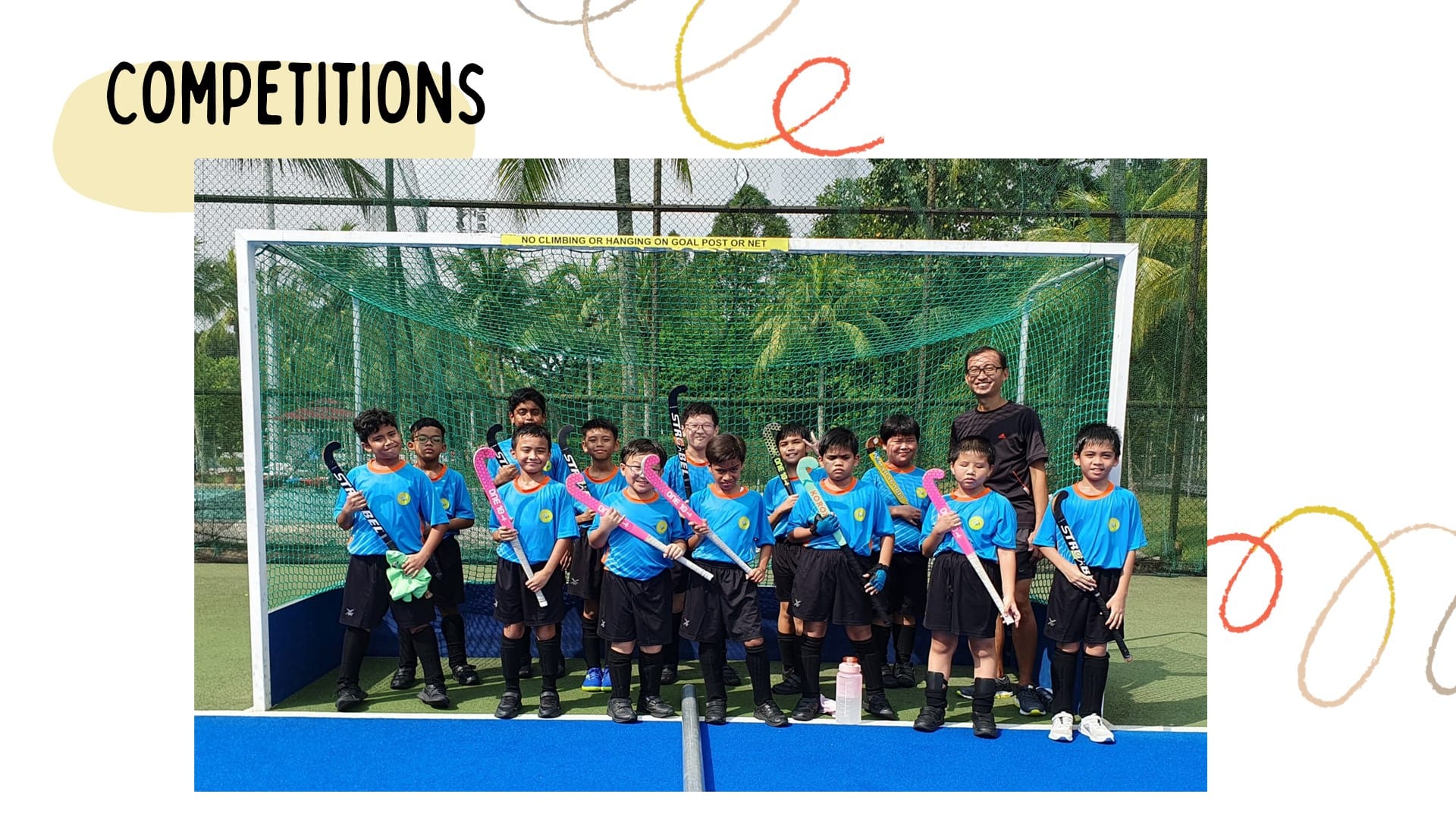 Young field hockey team in blue jerseys holding sticks, posing in front of a goal net with a coach.