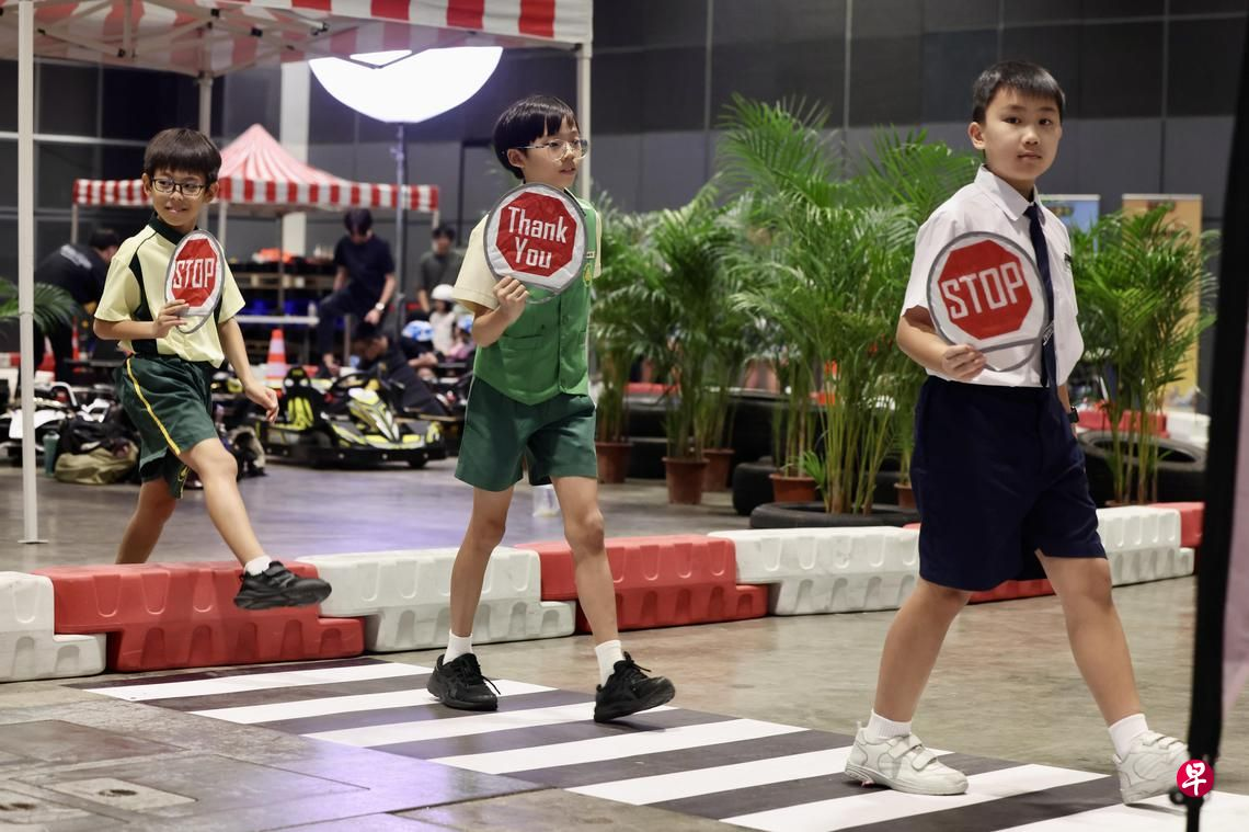 Three children in school uniforms hold stop signs while walking on a painted pedestrian crossing indoors.
