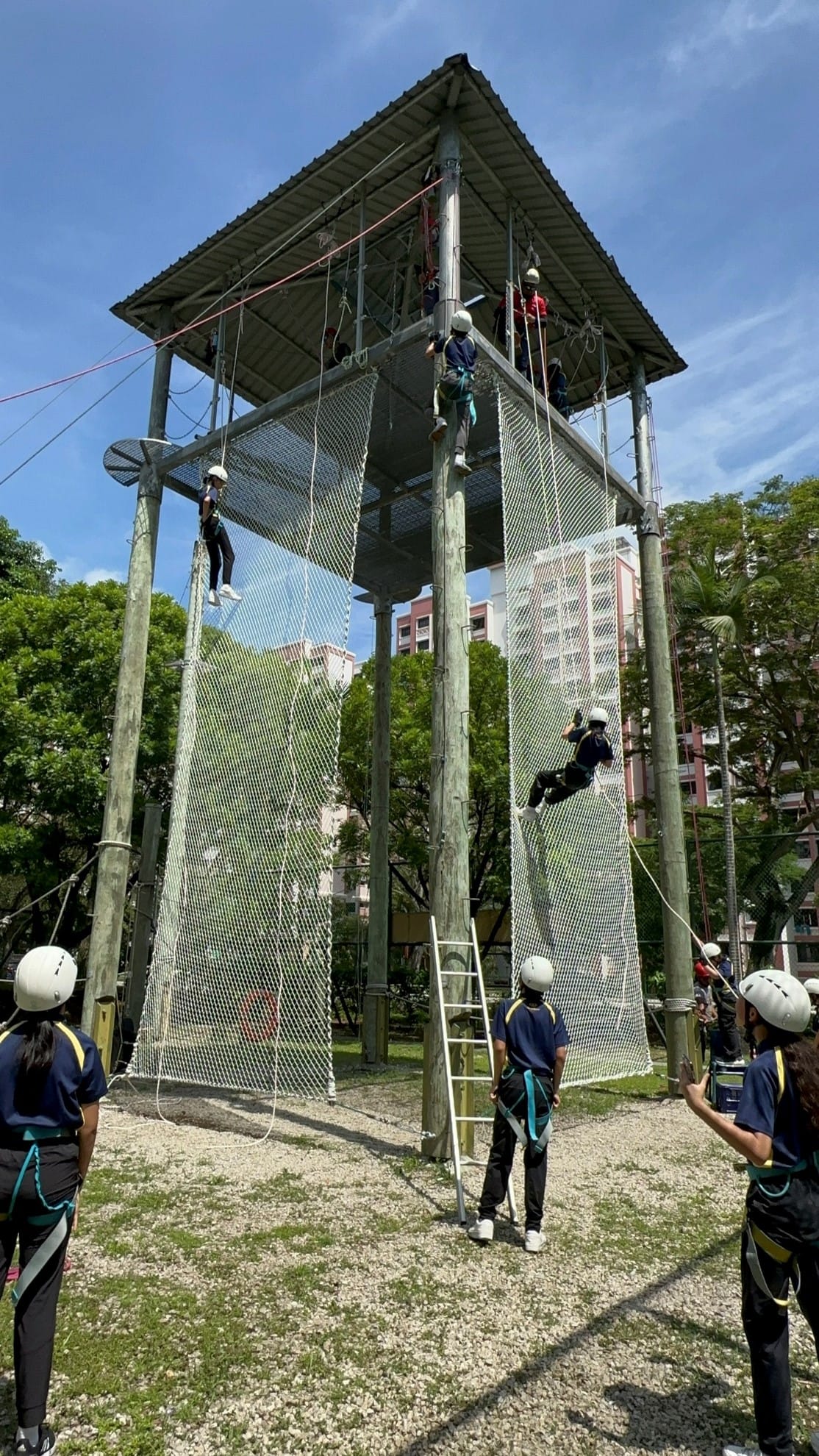 Students in helmets and harnesses climb nets and ropes on a tall outdoor adventure tower.