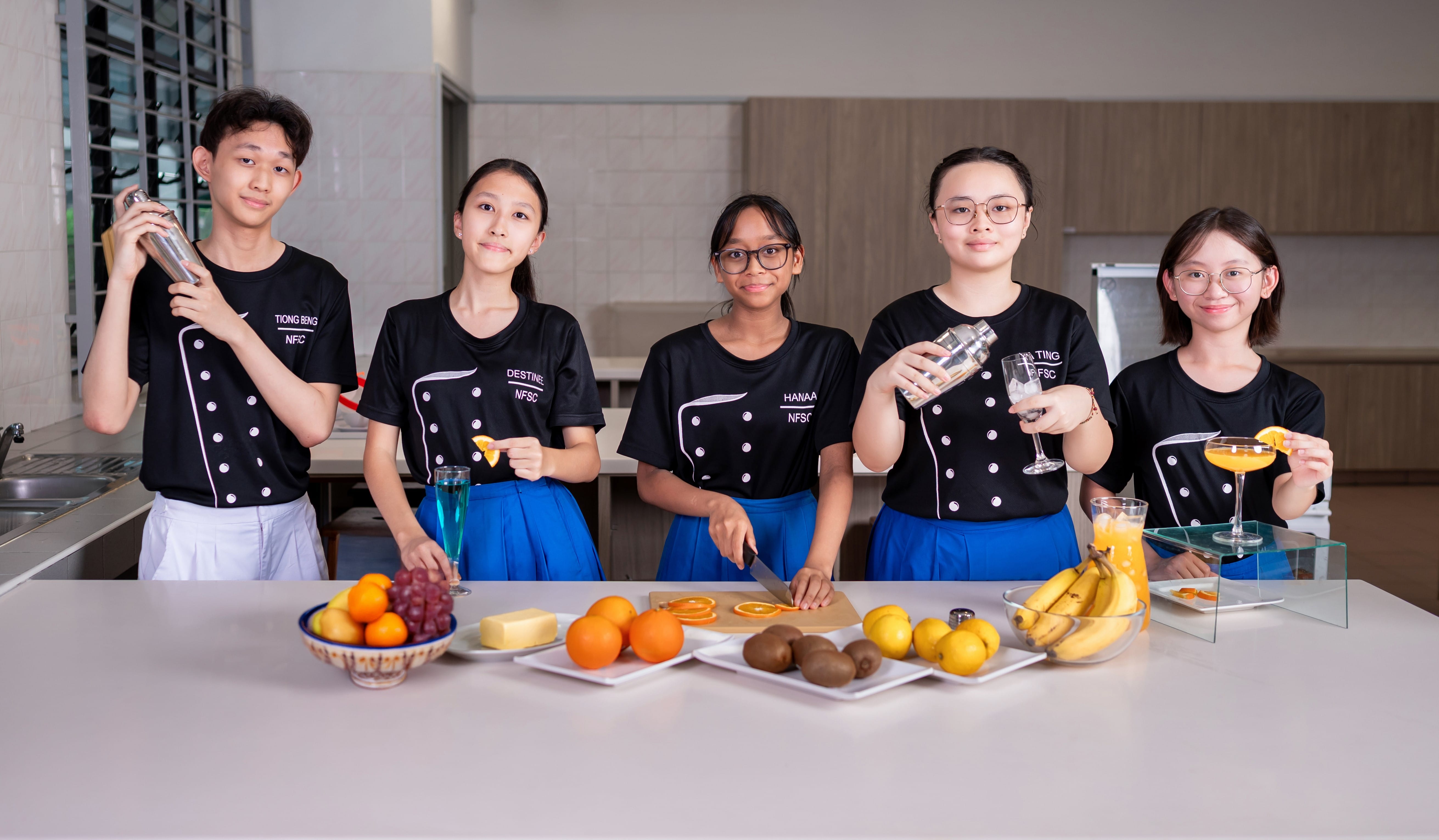 Five students in black NFSC uniforms stand behind a counter with fruits, cutting oranges and holding cocktail shakers.