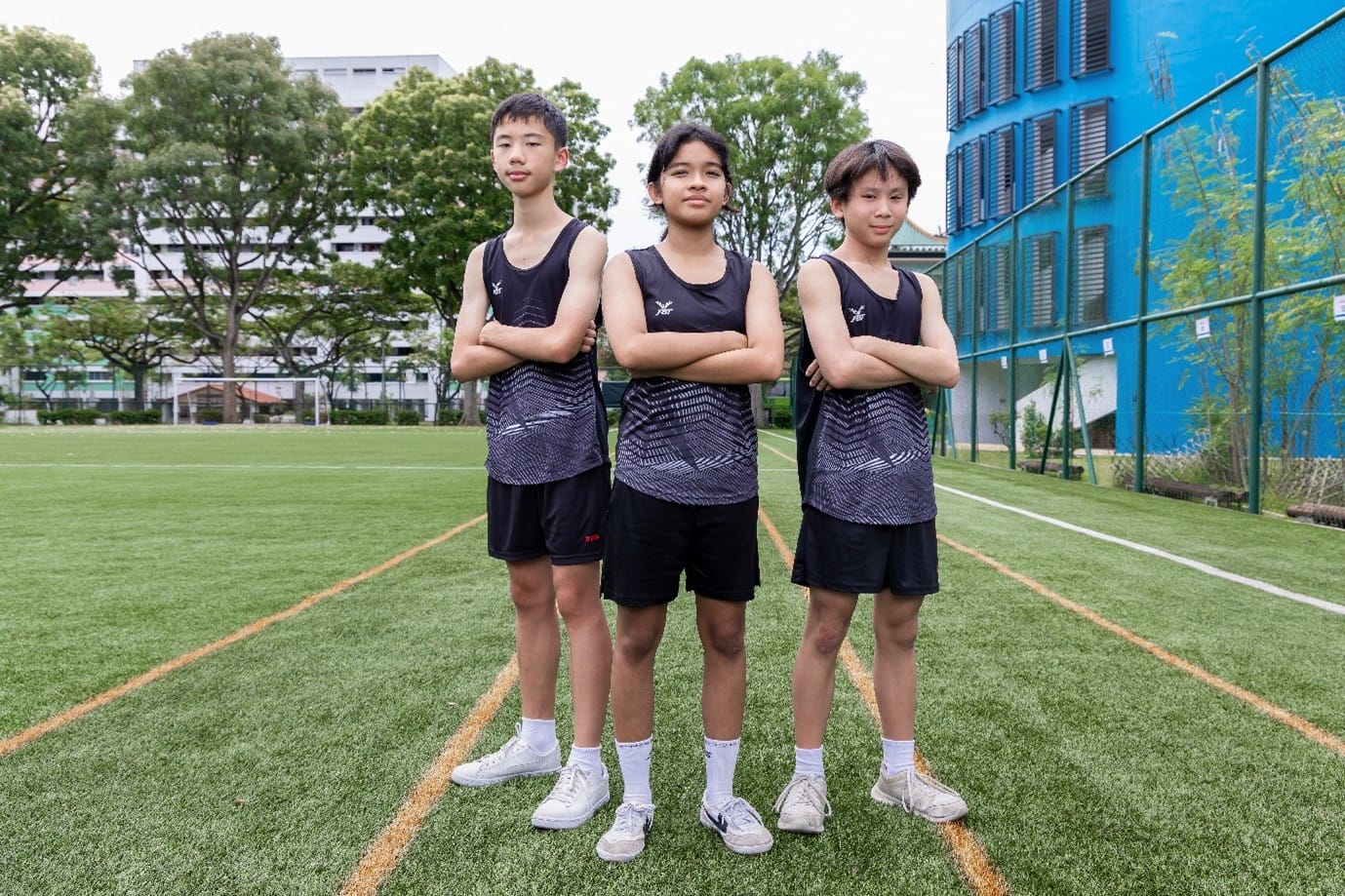 Three young athletes in matching black sleeveless uniforms stand with arms crossed on a green sports field.
