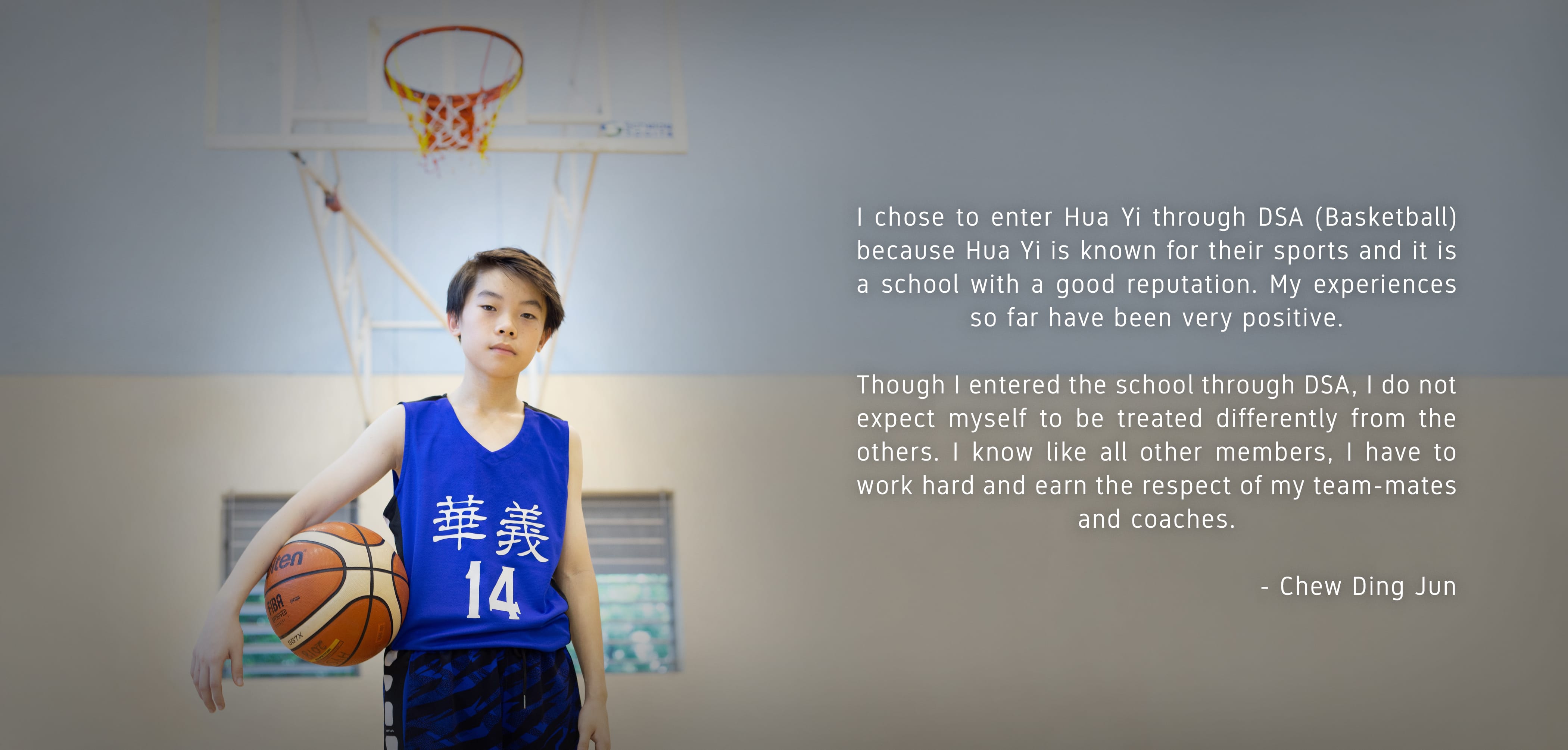Young boy in blue jersey number 14 holds a basketball in a gym with a hoop visible behind him.