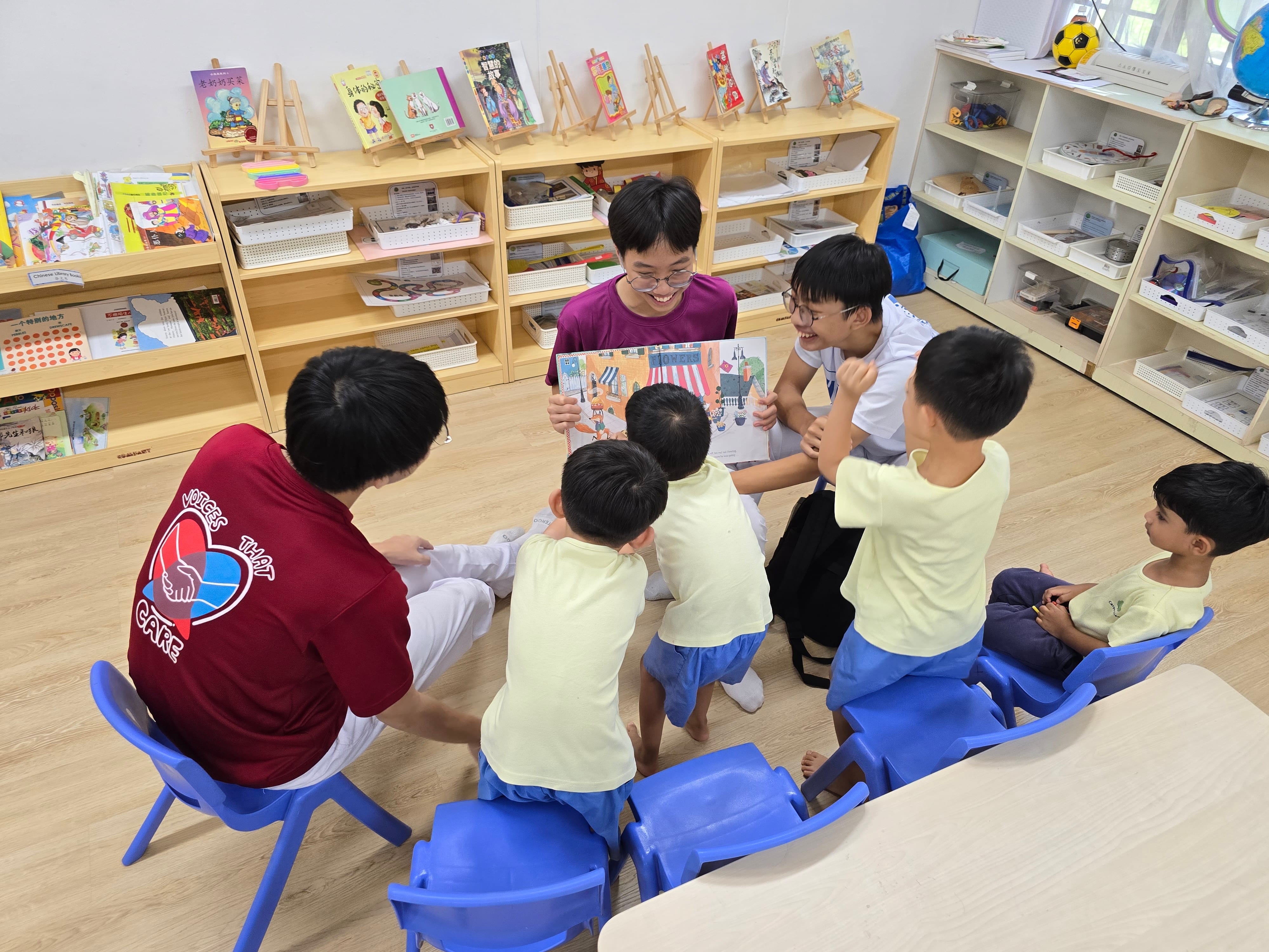 Two teenagers read a picture book to four young children sitting on blue chairs in a classroom with bookshelves.
