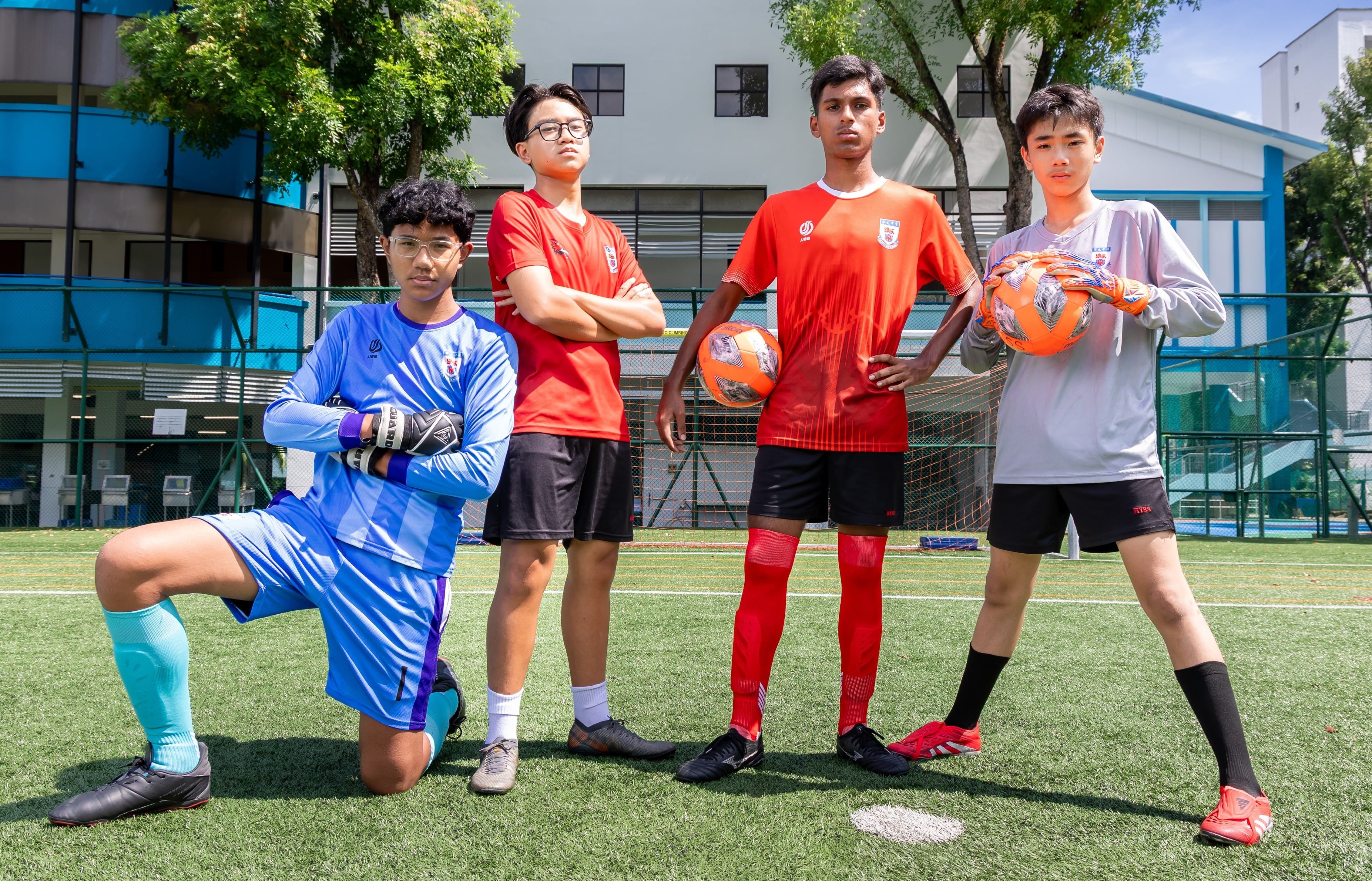 Four football players, including two goalkeepers holding balls, pose on a grass pitch in team kits.