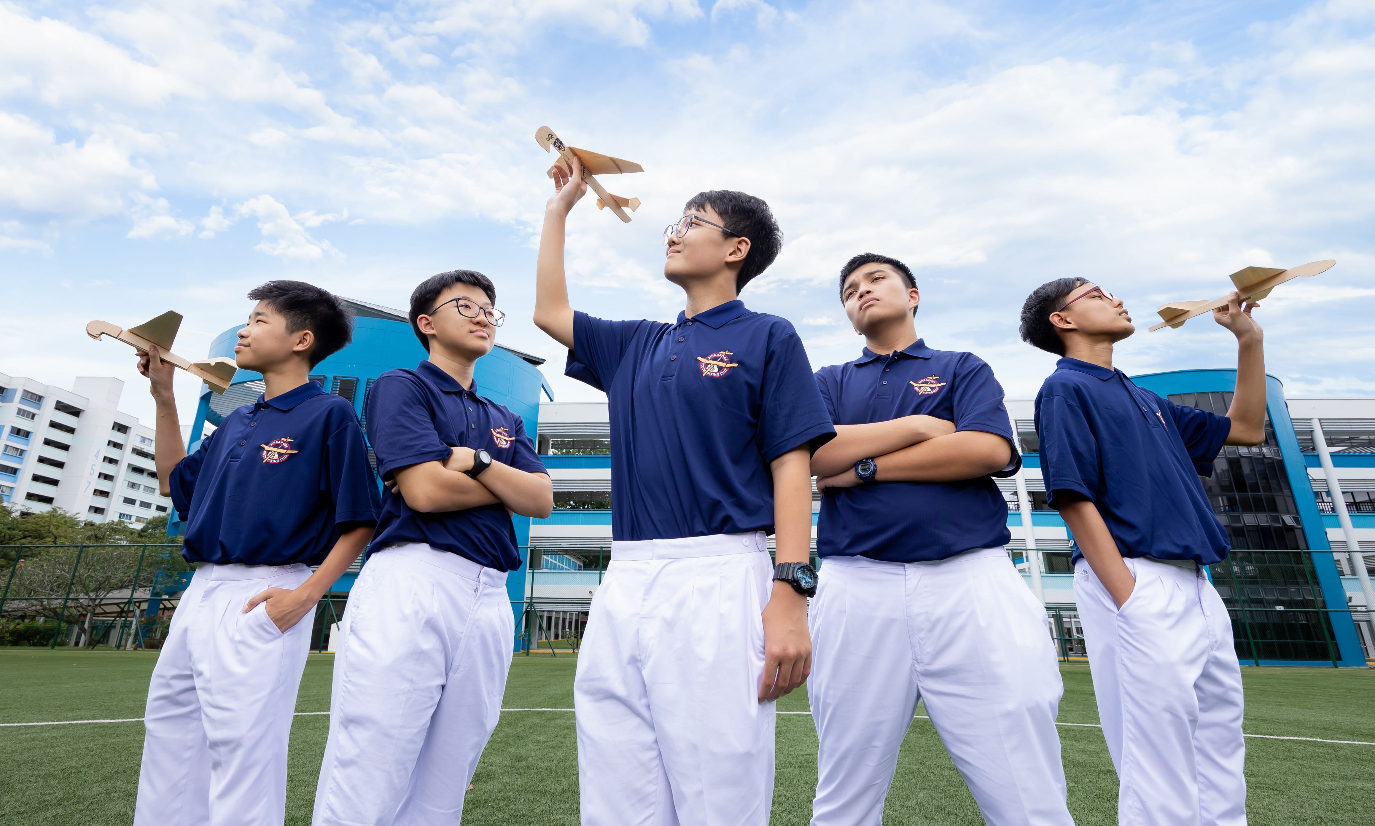 Five students in navy and white uniforms stand on a field holding wooden model planes toward a cloudy sky.