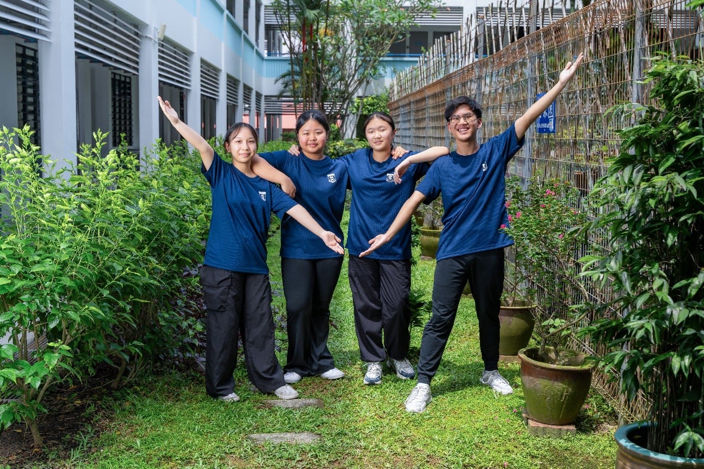 Four students in matching blue shirts and black pants pose with arms outstretched in a green school garden.