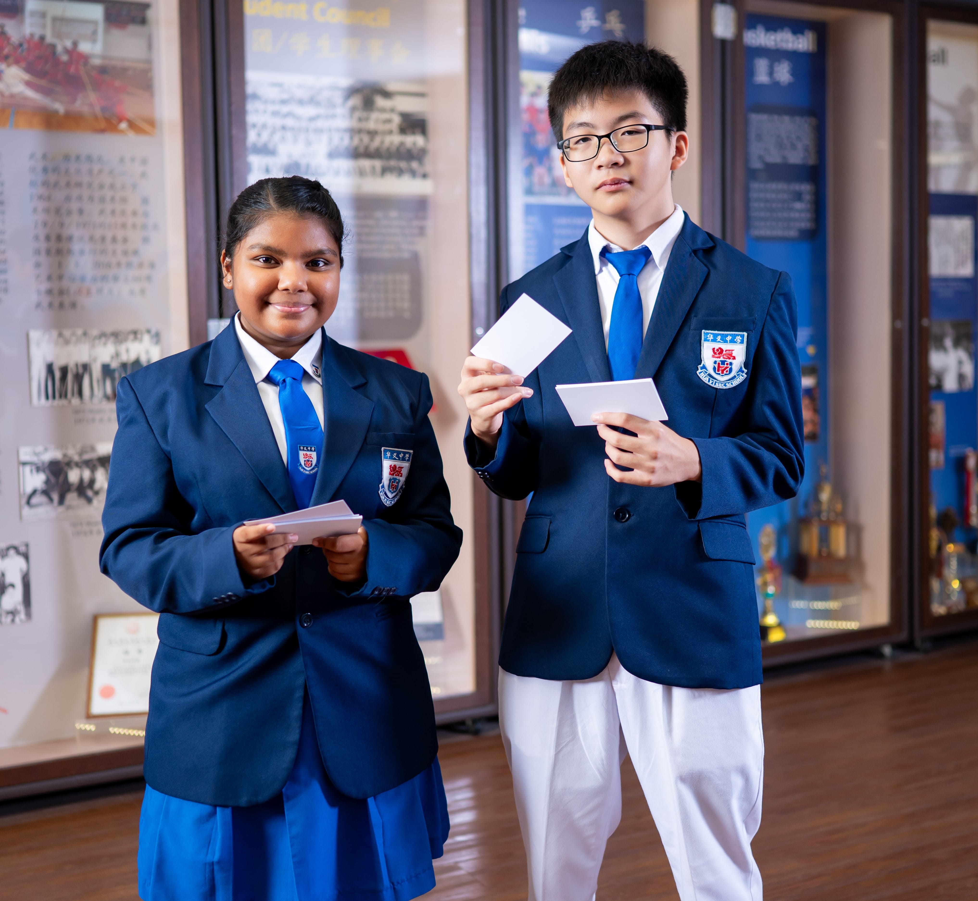 Two students in navy blazers and blue ties hold cards, standing before school display boards.