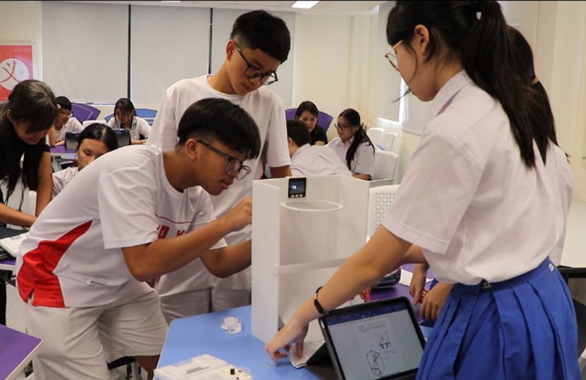 Three students in white uniforms work on a project with a tablet and white structure in a modern classroom.