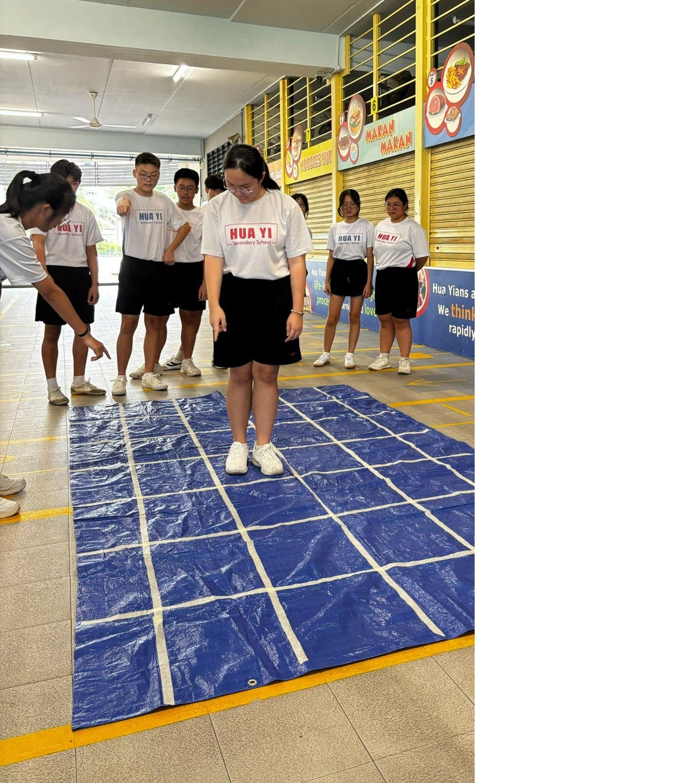 Students in Hua Yi Secondary School uniforms stand around a blue tarp grid on a covered walkway floor.