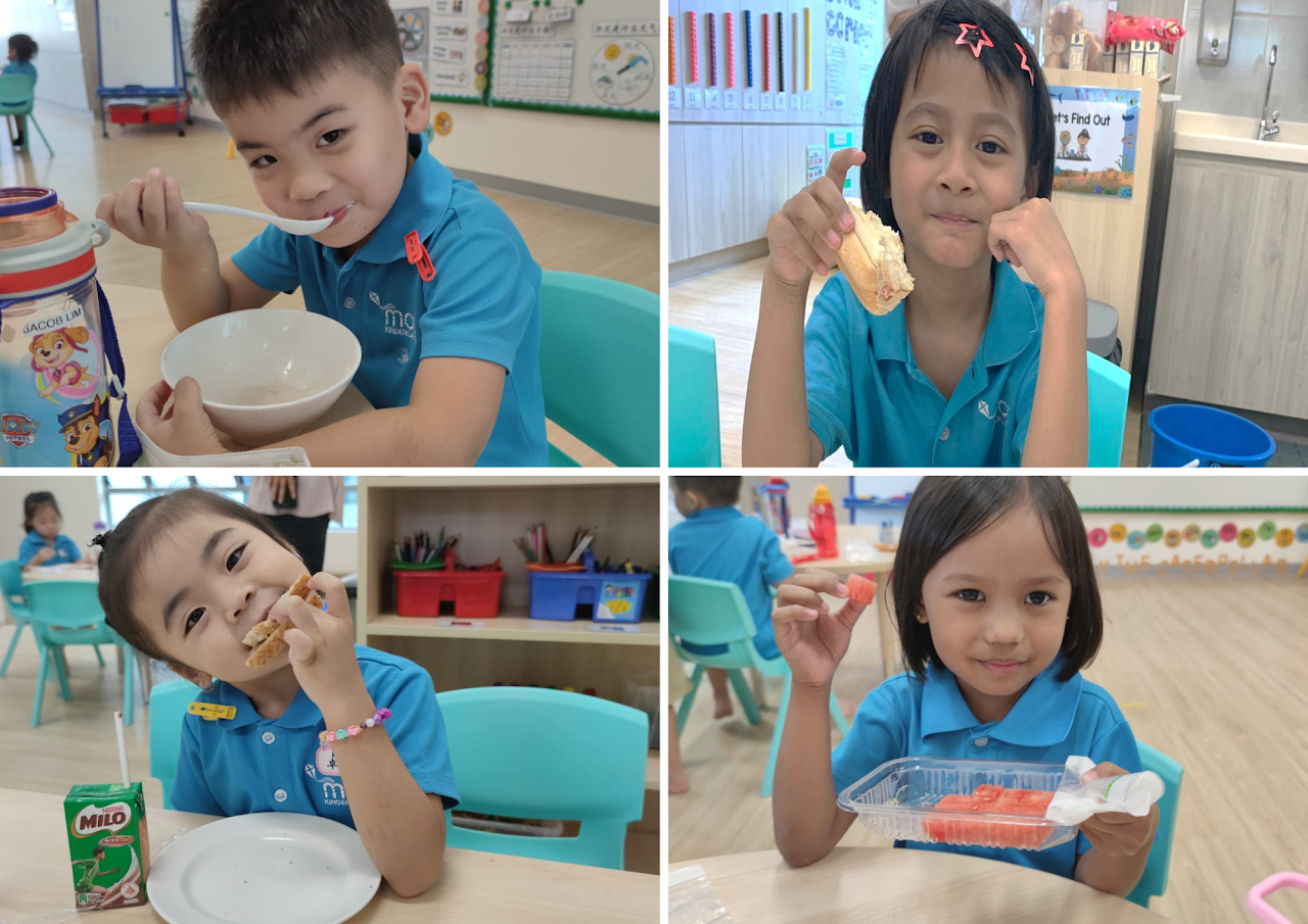 Four children in blue uniforms sit at classroom tables, smiling while eating soup, bread, and watermelon.