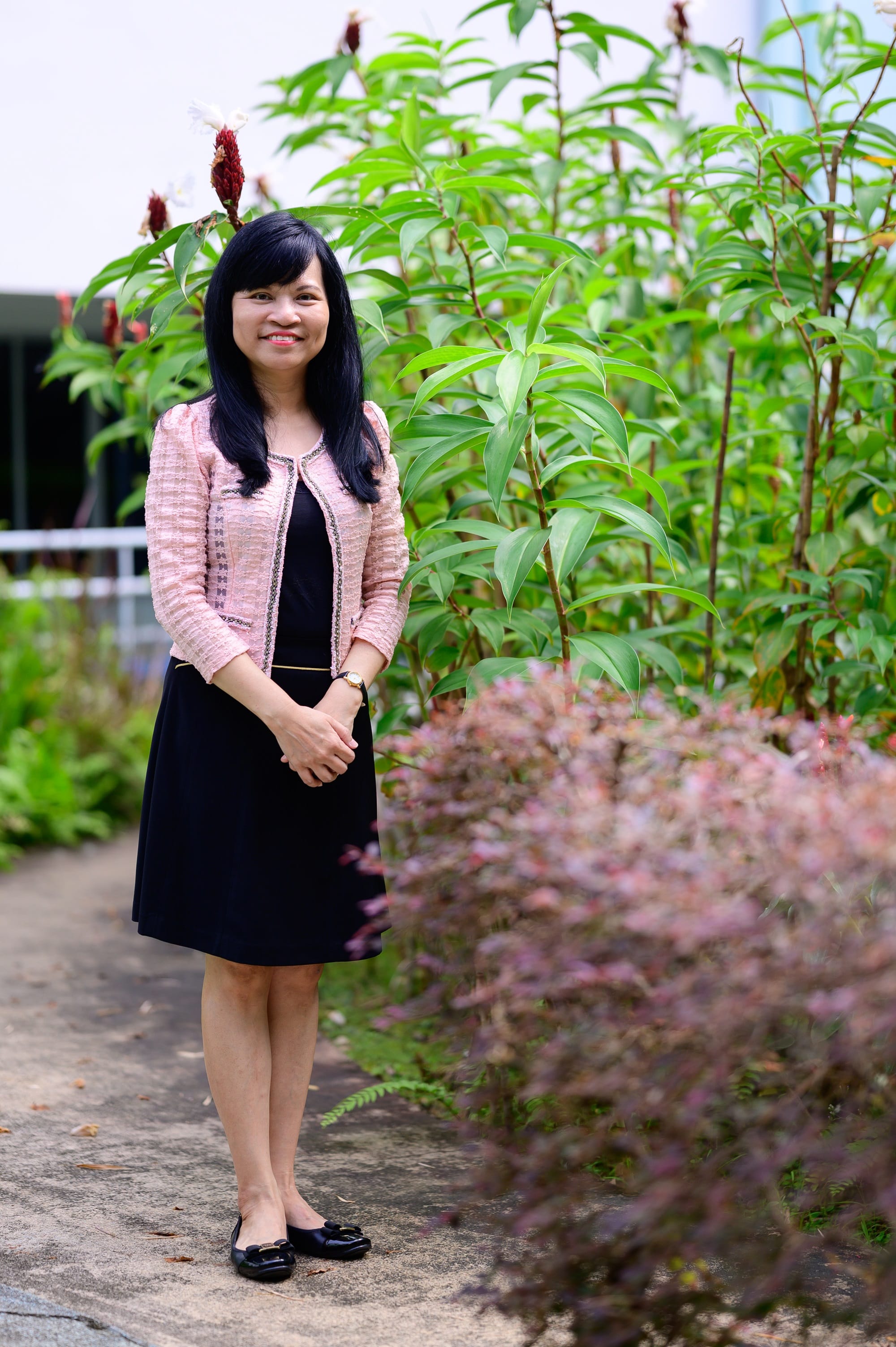 A smiling woman in a black dress and pink jacket stands on a path surrounded by green and pink foliage.
