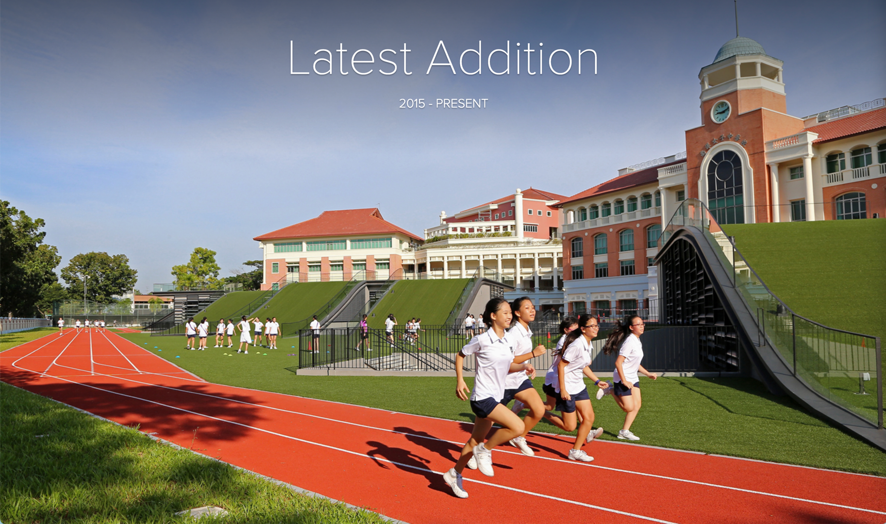 Students in uniforms running on a red track beside a green hill structure and red-roofed school buildings.