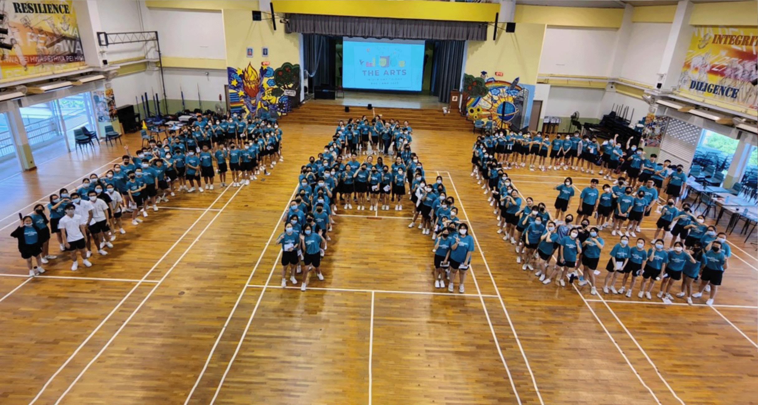 Students in teal shirts form a large number shape on a school hall floor, with a stage and "The Arts" screen behind them.