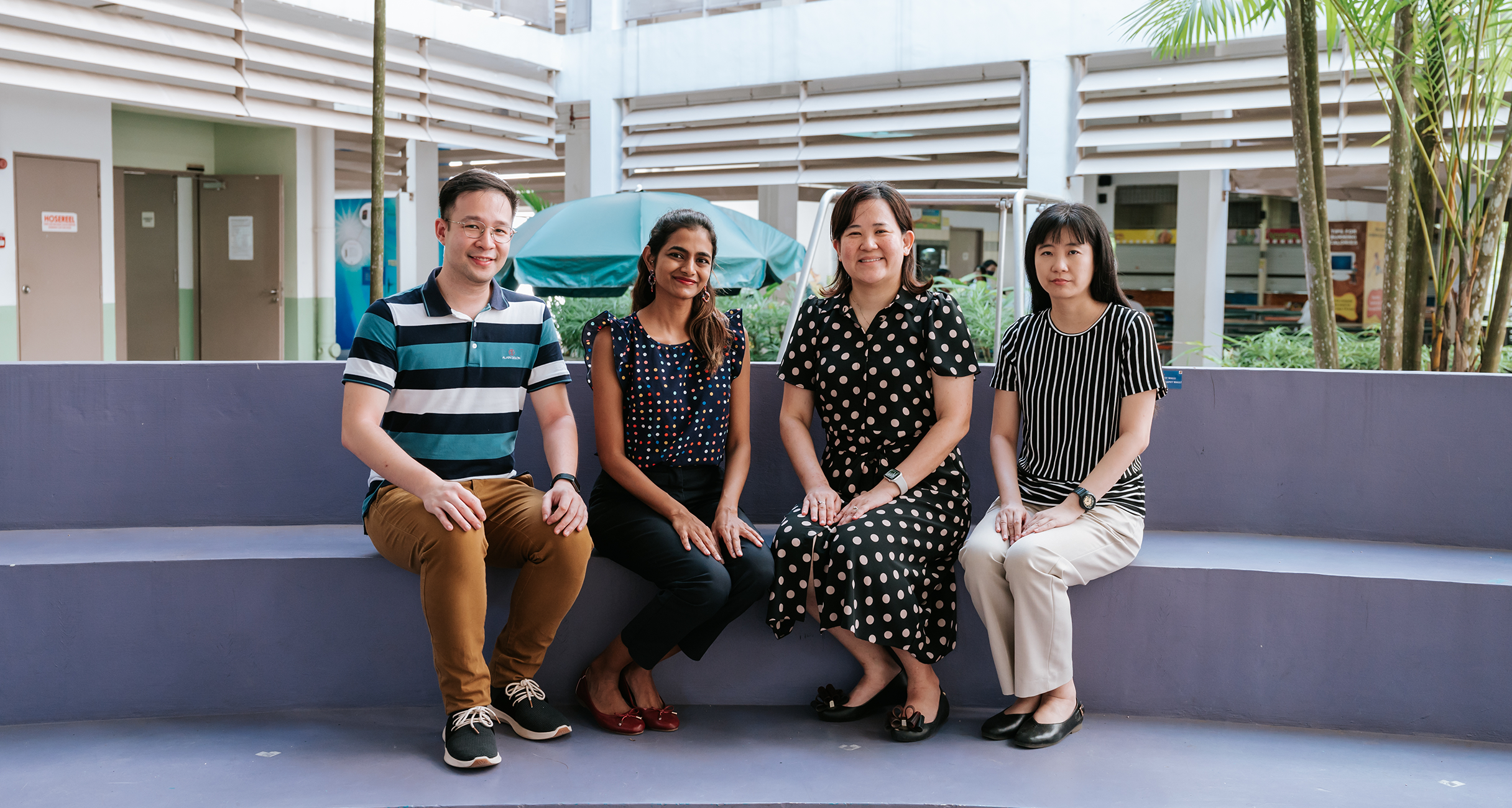 Four people sit on a curved bench in an open-air courtyard, posing and smiling for a group photo.