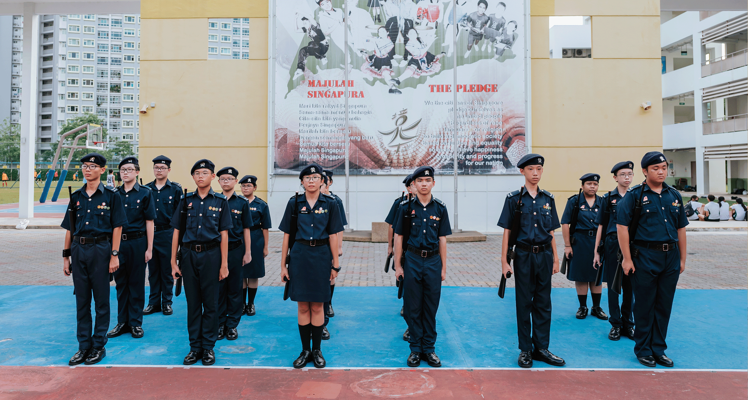Young students in dark navy uniforms and berets stand at attention on a court in front of a Majulah Singapura mural.
