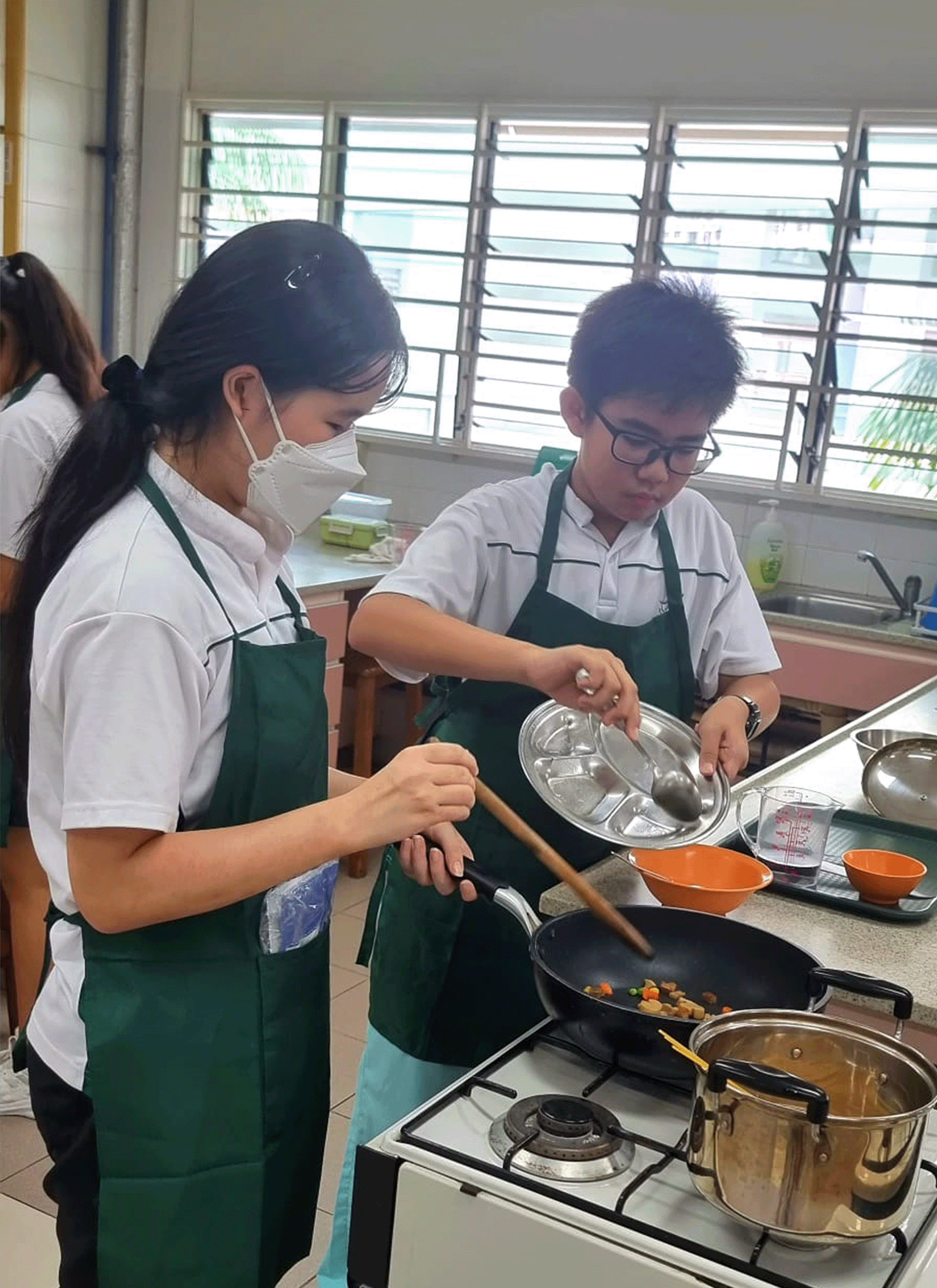 Two students in green aprons cook vegetables in a pan on a gas stove during a school cooking class.
