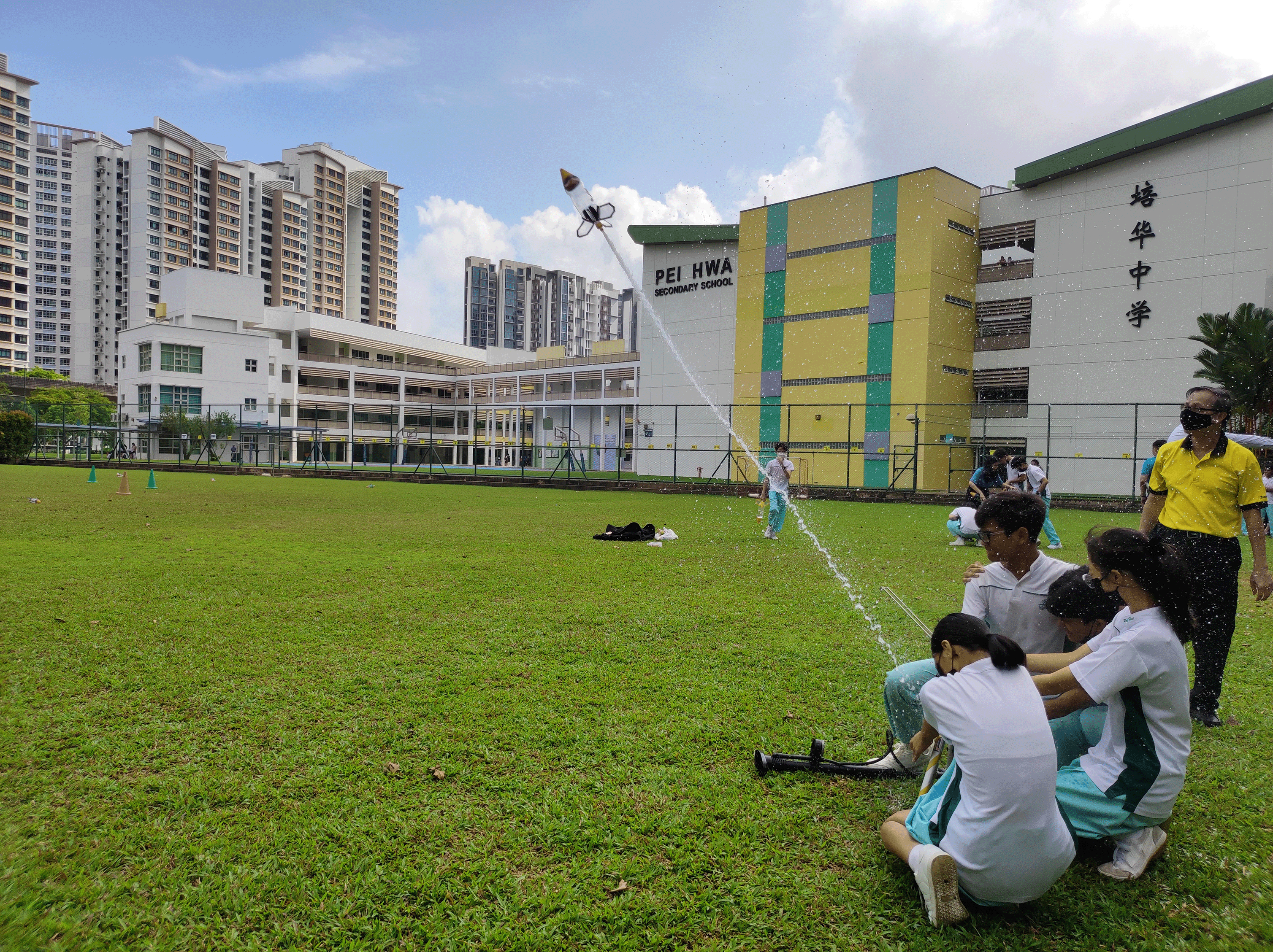 Students on a grassy field launch a model rocket with a white smoke trail beside a school building.