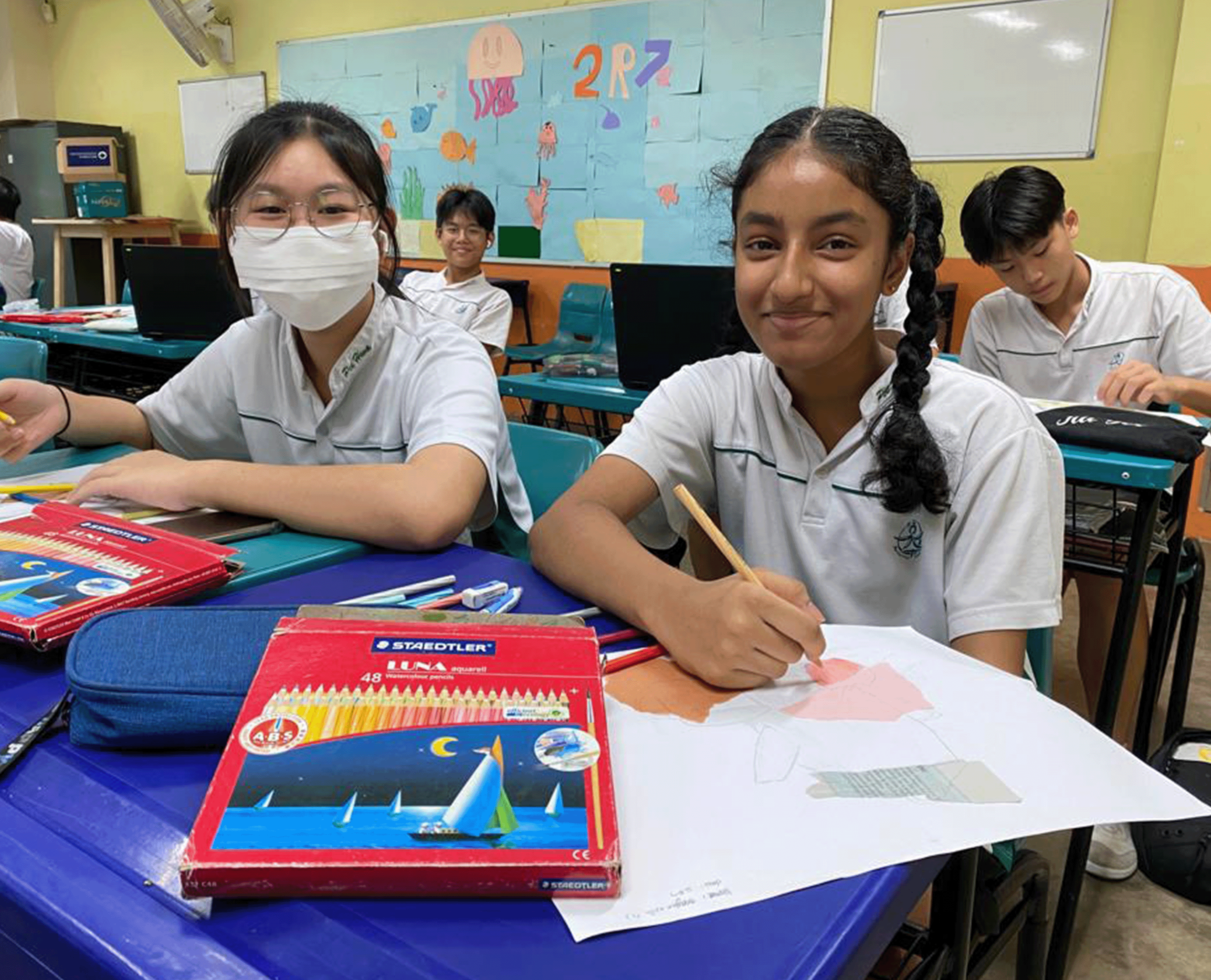 Four students in white uniforms sit at desks in a colorful classroom, one girl drawing with colored pencils.
