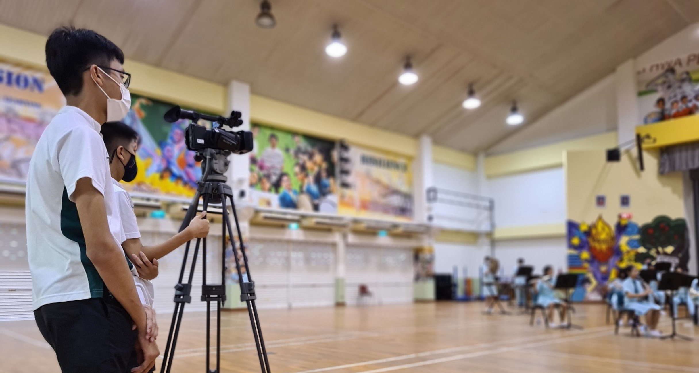 Two masked students stand beside a video camera on a tripod filming a group seated in a school hall.
