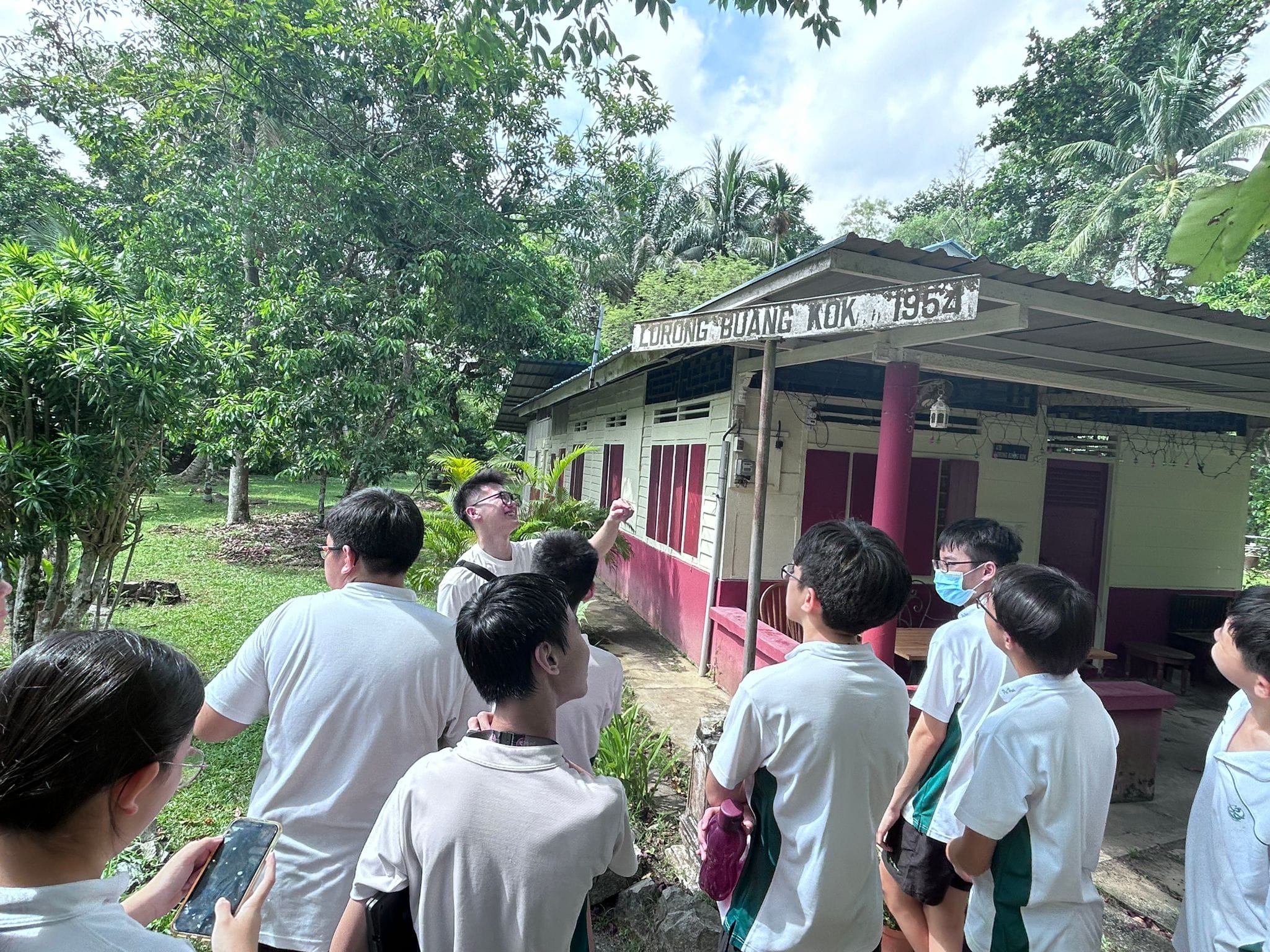 Students in white shirts gather outside an old building signed Lorong Buang Kok 1962, surrounded by trees.