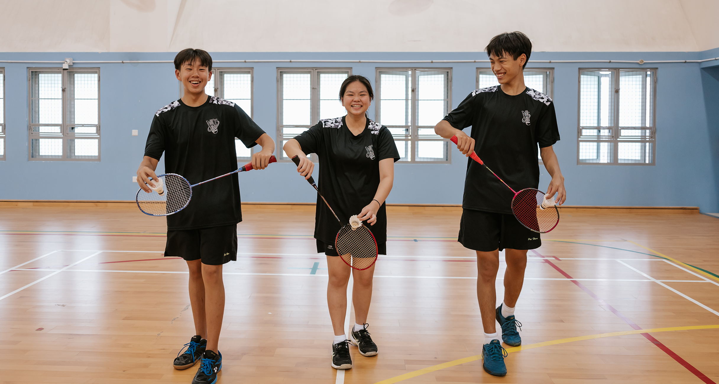 Three smiling teenagers in black uniforms hold badminton rackets and shuttlecocks on an indoor wooden court.