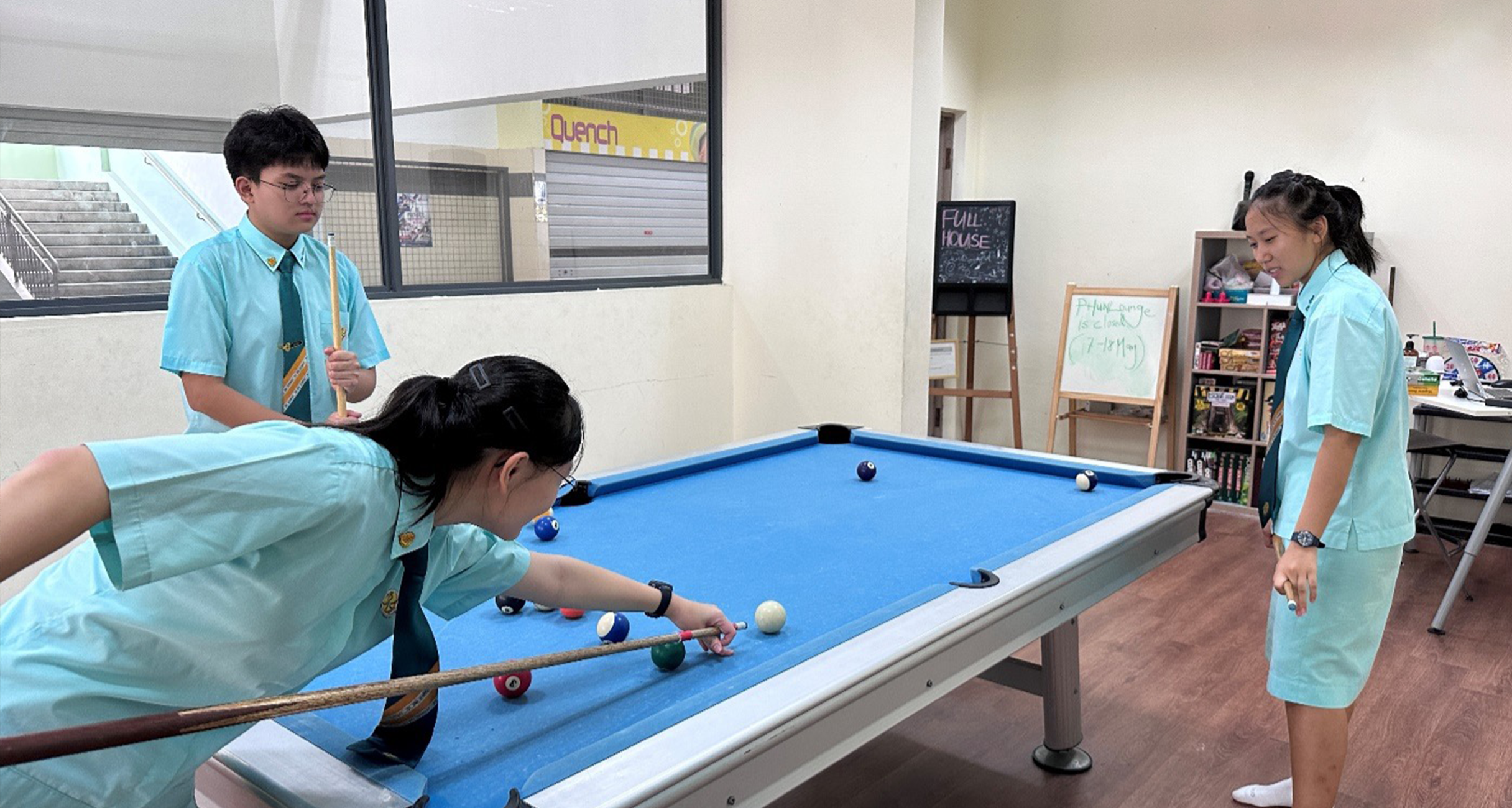 Three students in light blue uniforms playing pool on a blue billiard table indoors.