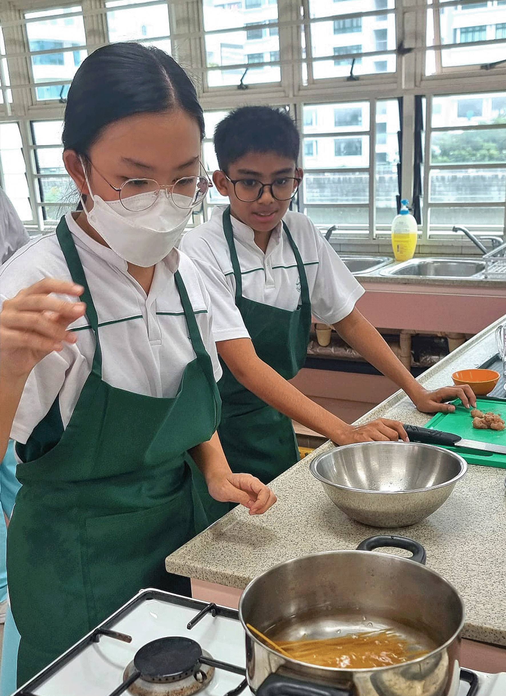Two students in green aprons cook in a school kitchen, with a pot on a gas stove and a mixing bowl on the counter.
