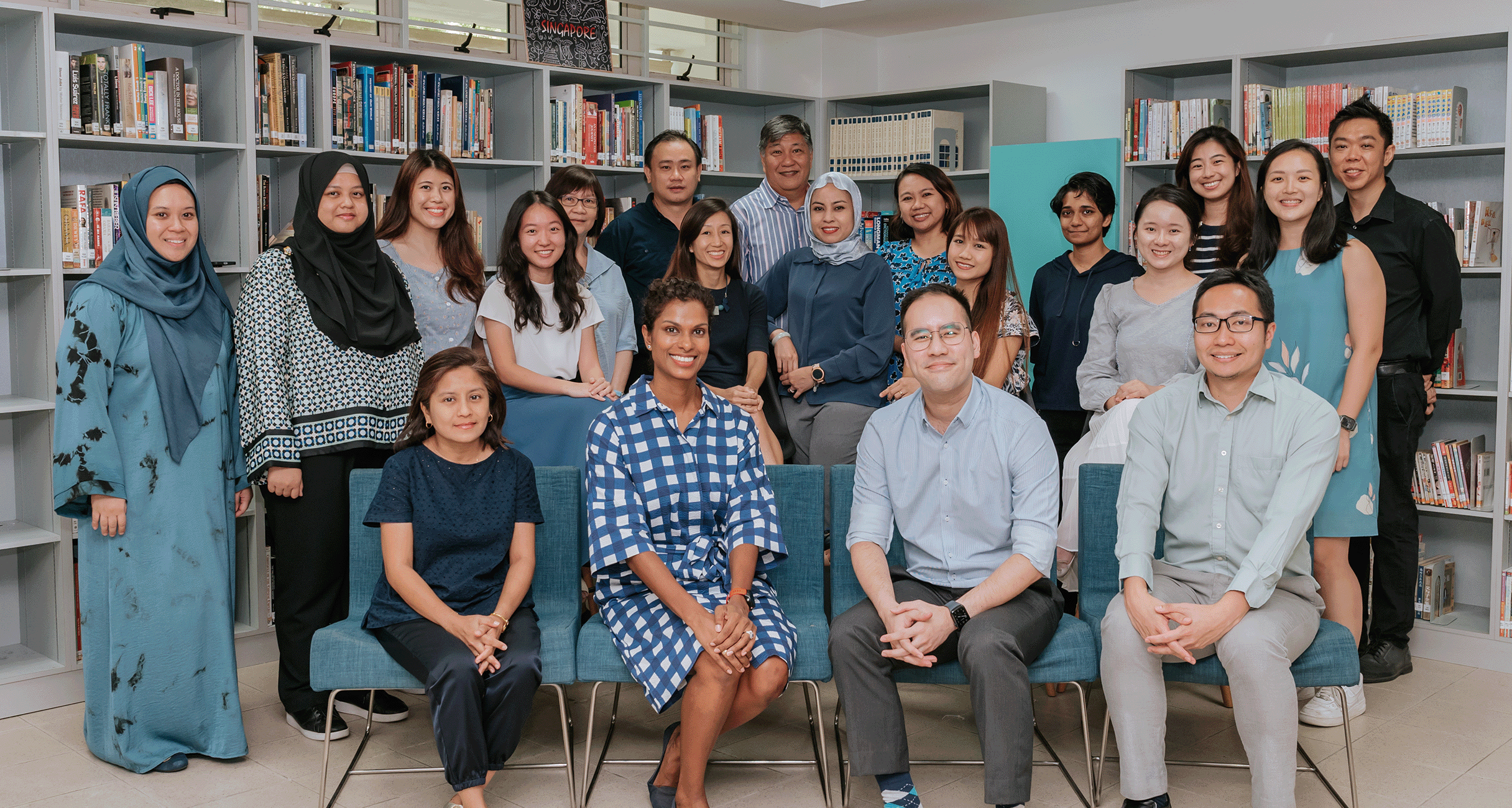 A large diverse group of about 20 people smiling together in a library with bookshelves in the background.