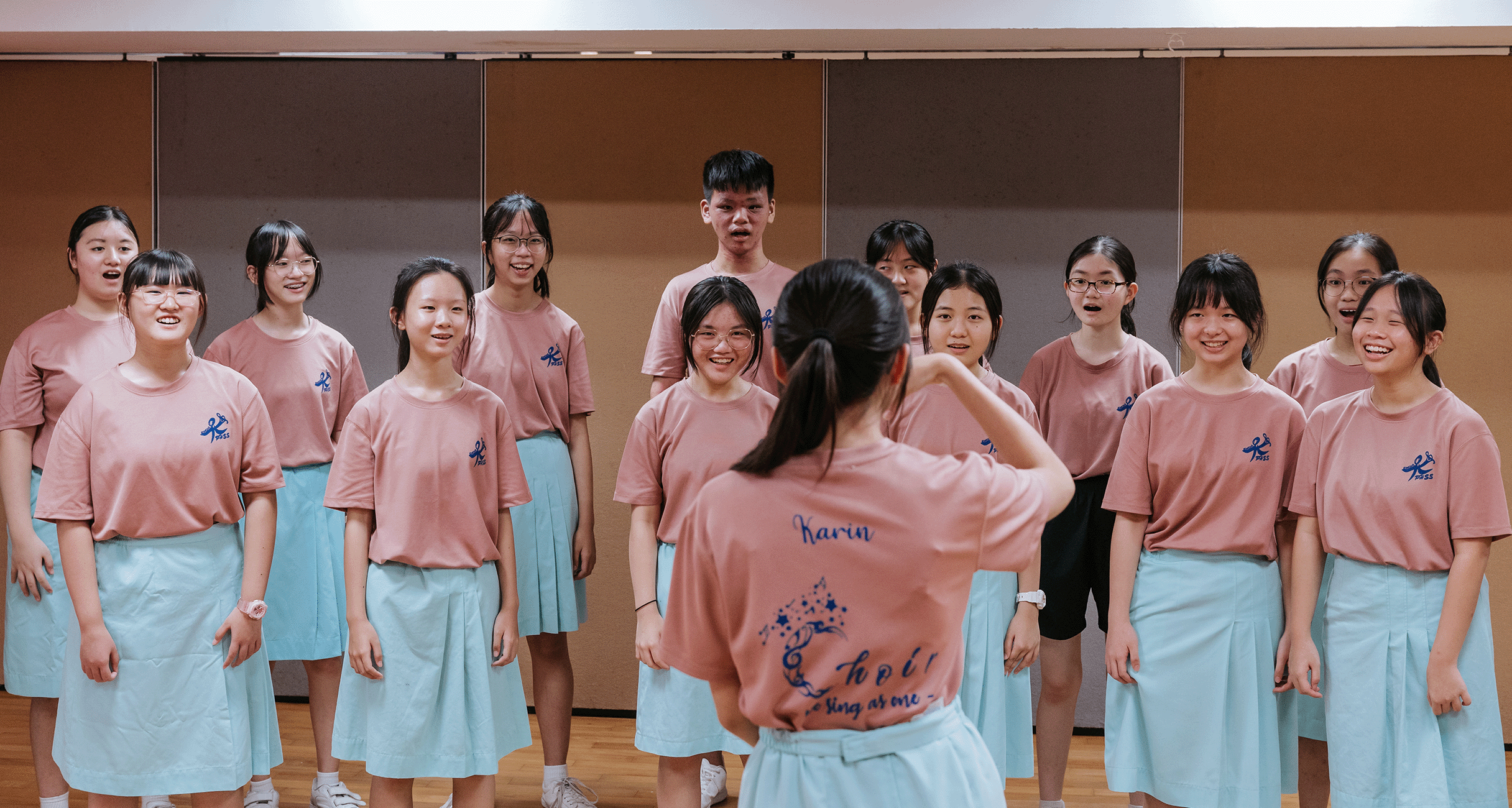 A student conductor named Karin leads a youth choir in pink shirts and light blue skirts during rehearsal.