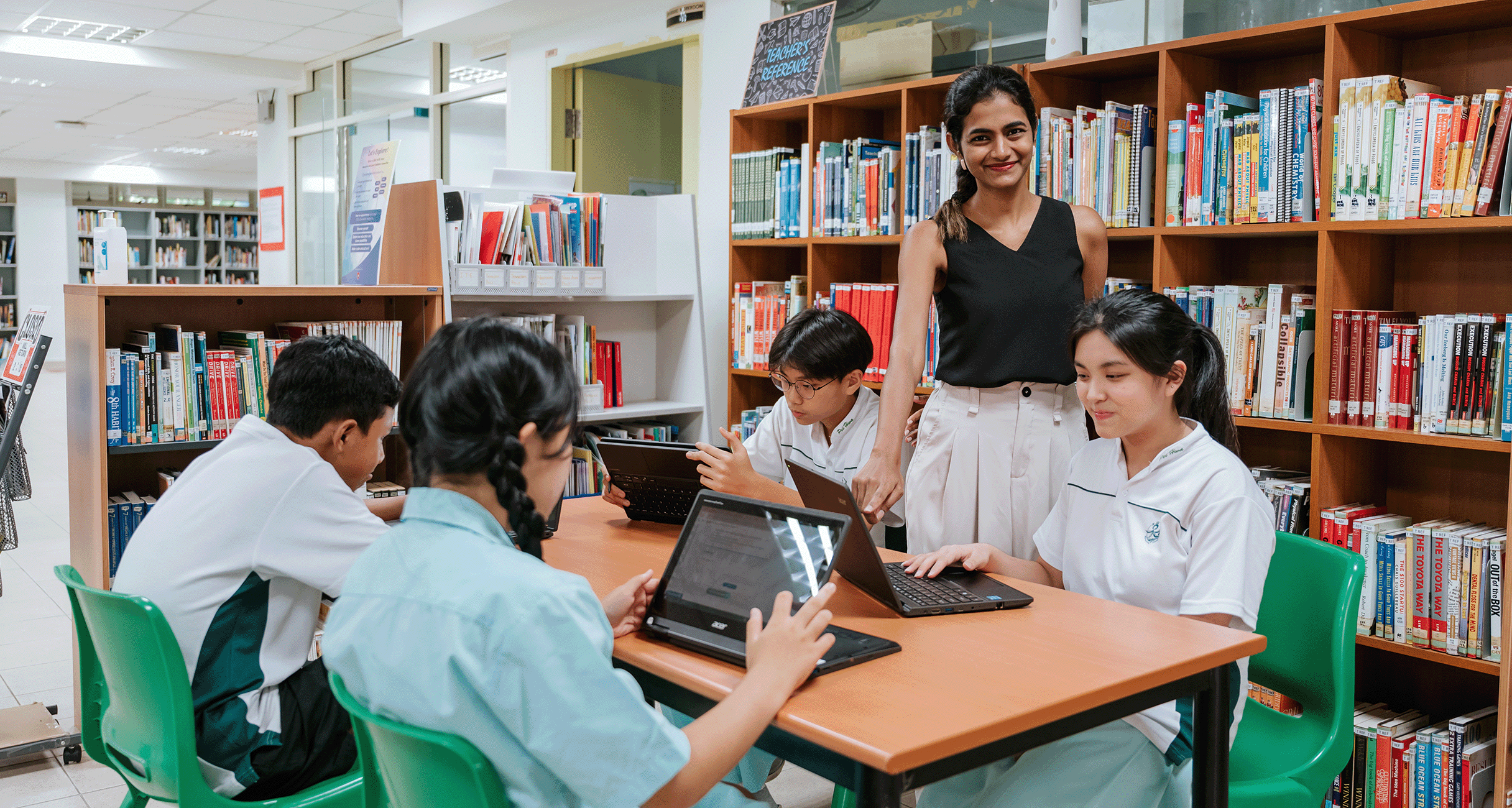 A smiling teacher stands over four students using laptops at a table in a school library with bookshelves behind them.