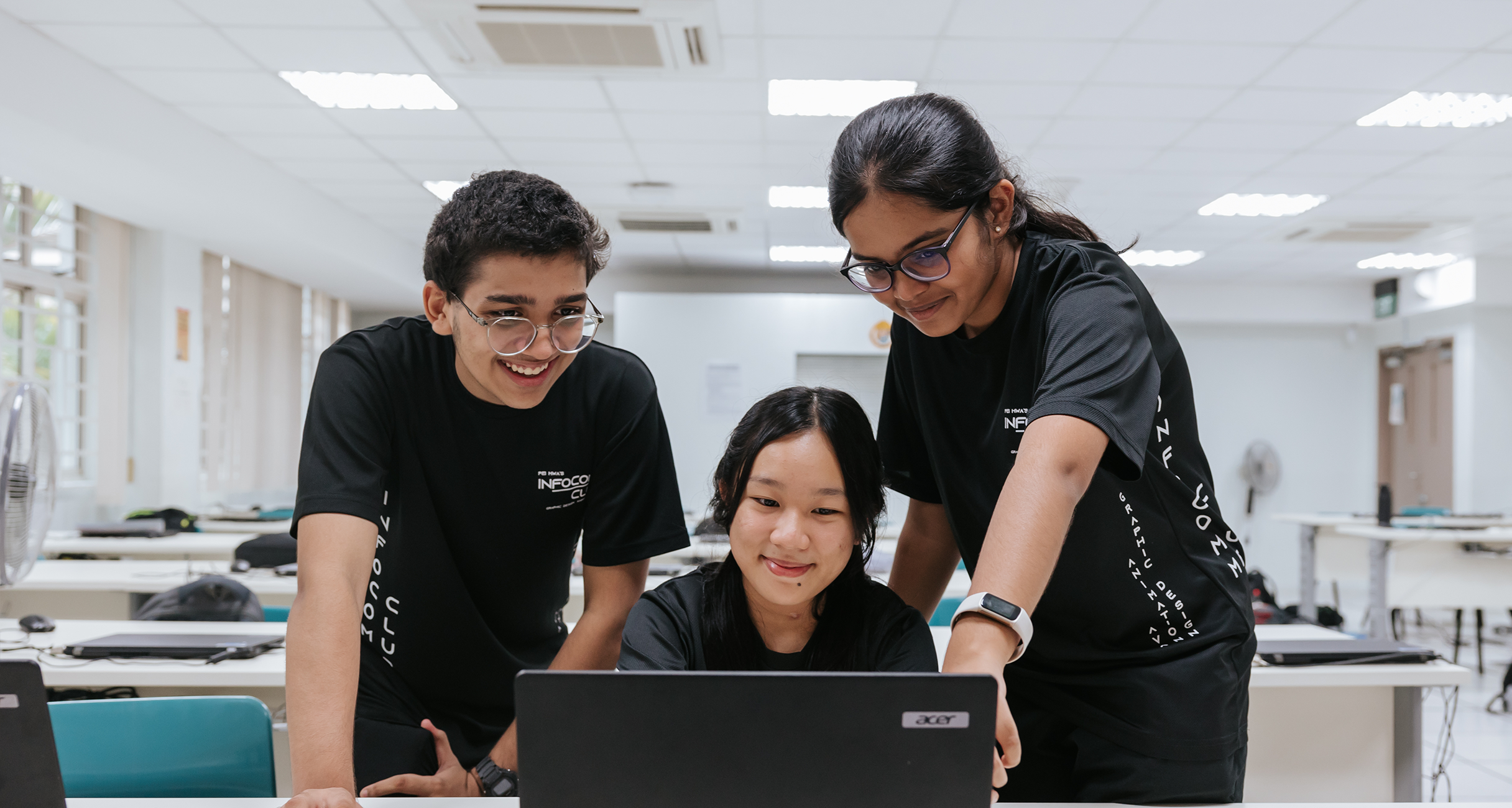 Three teenagers in black shirts smile while gathered around an Acer laptop in a bright classroom.