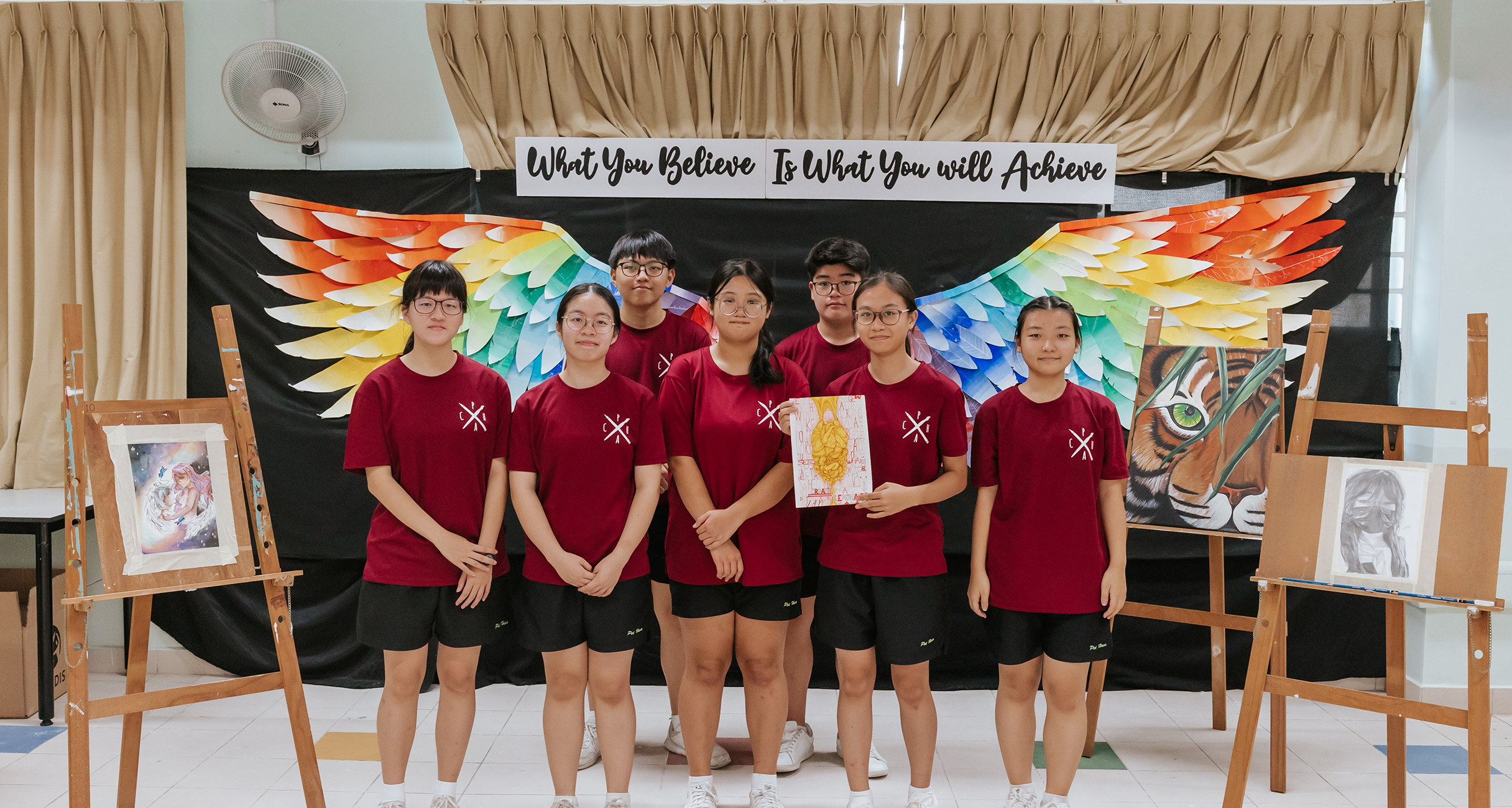 Seven students in maroon shirts pose before rainbow wings; artworks on easels flank them.