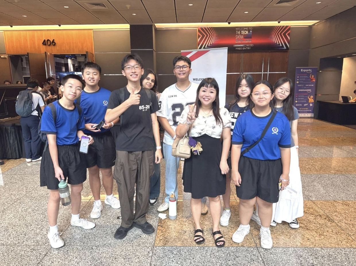 A group of nine students and an adult posing together inside a convention center near a Tech Show banner.