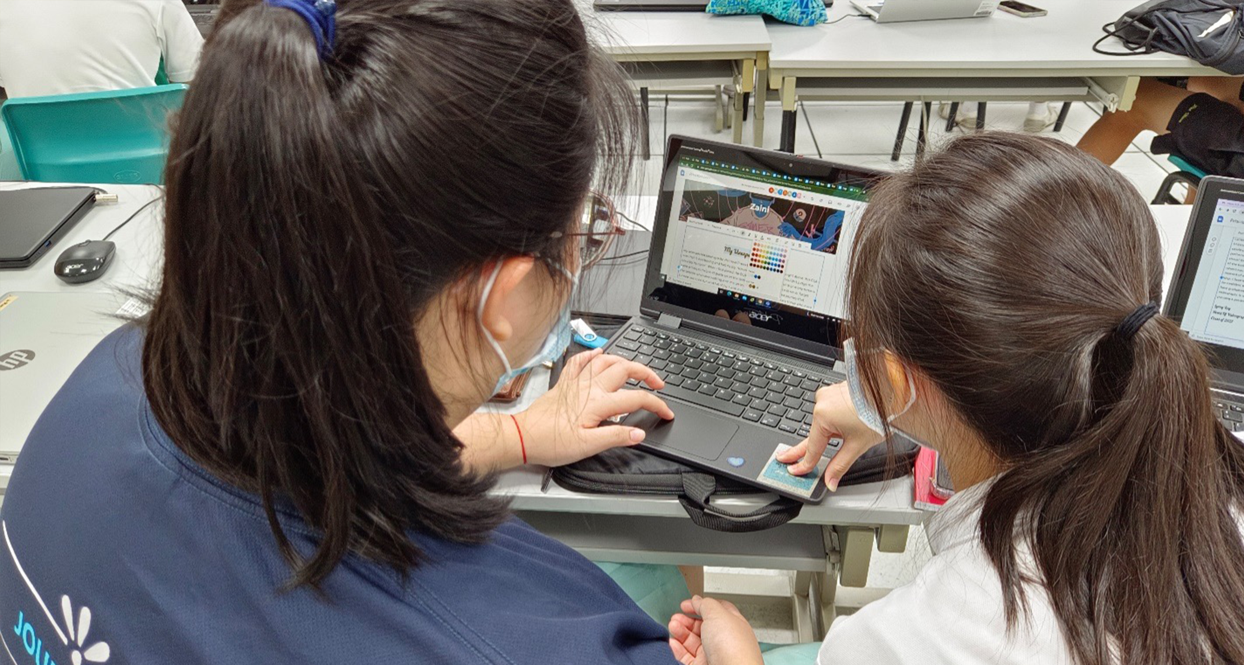 Two students with ponytails and masks work together on a laptop at a classroom desk.
