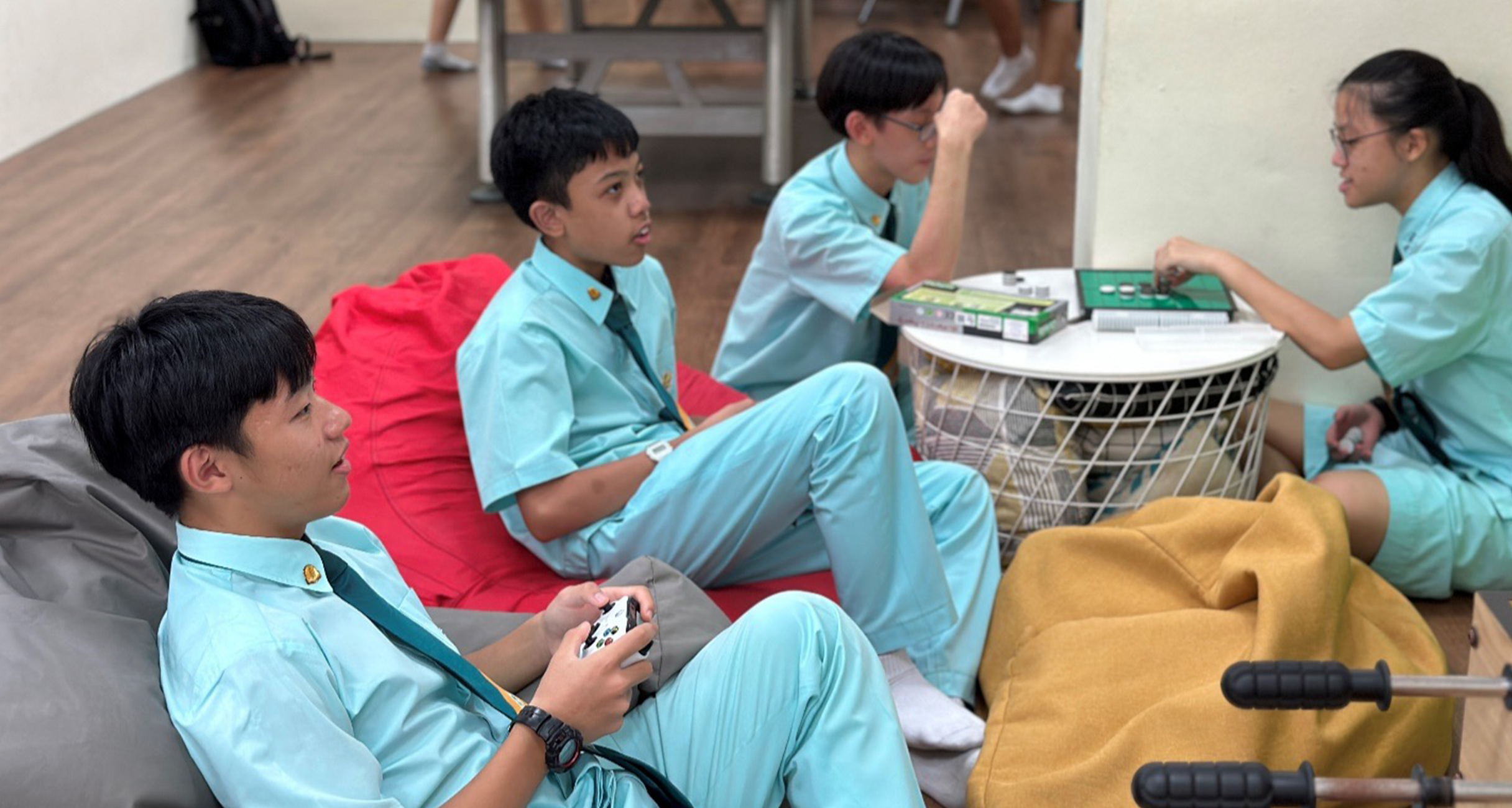 Four students in teal uniforms relax on bean bags; one holds a game controller while others play a board game.