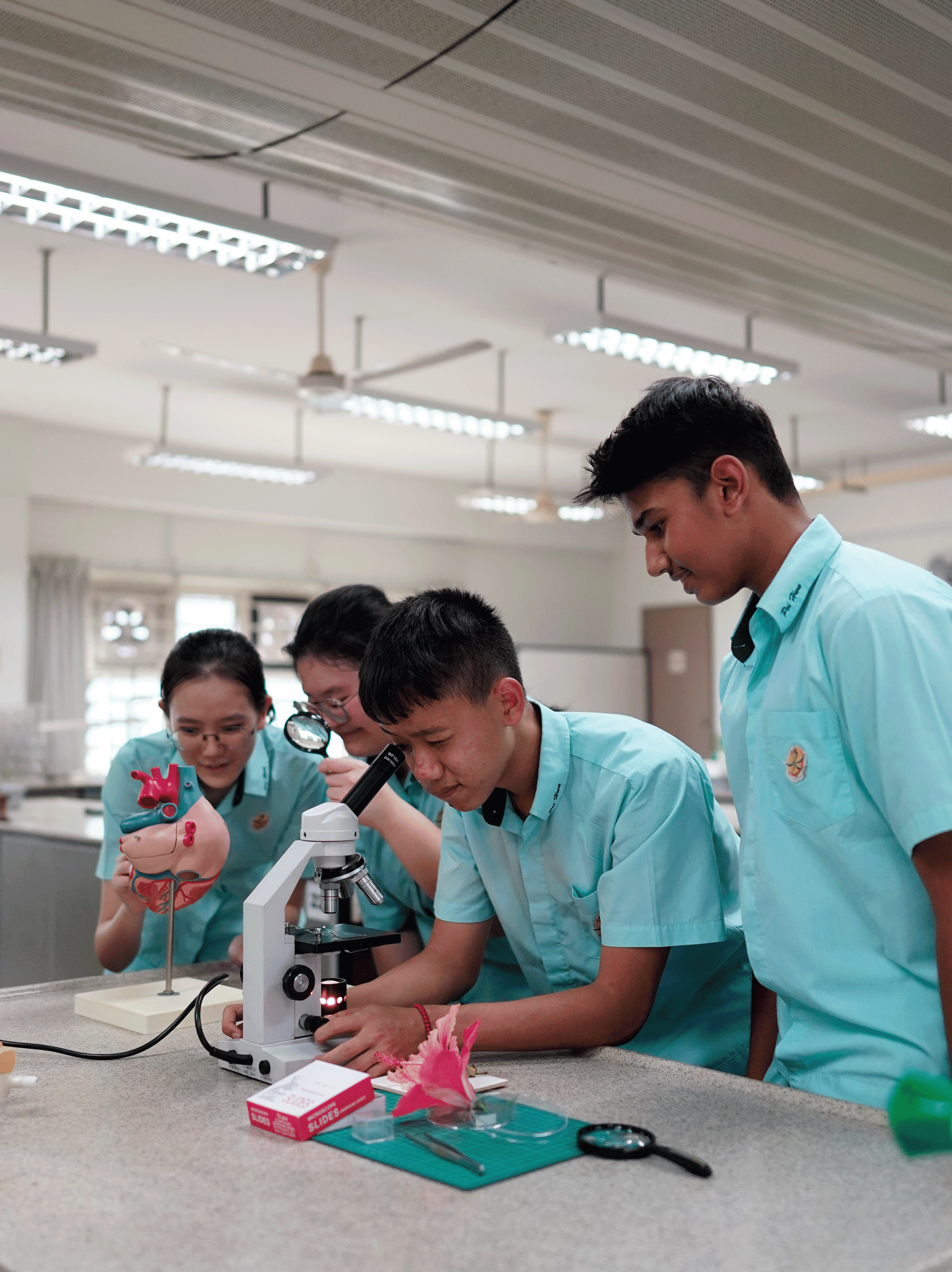 Four students in light blue uniforms examine a microscope on a lab bench with a heart model and tools.