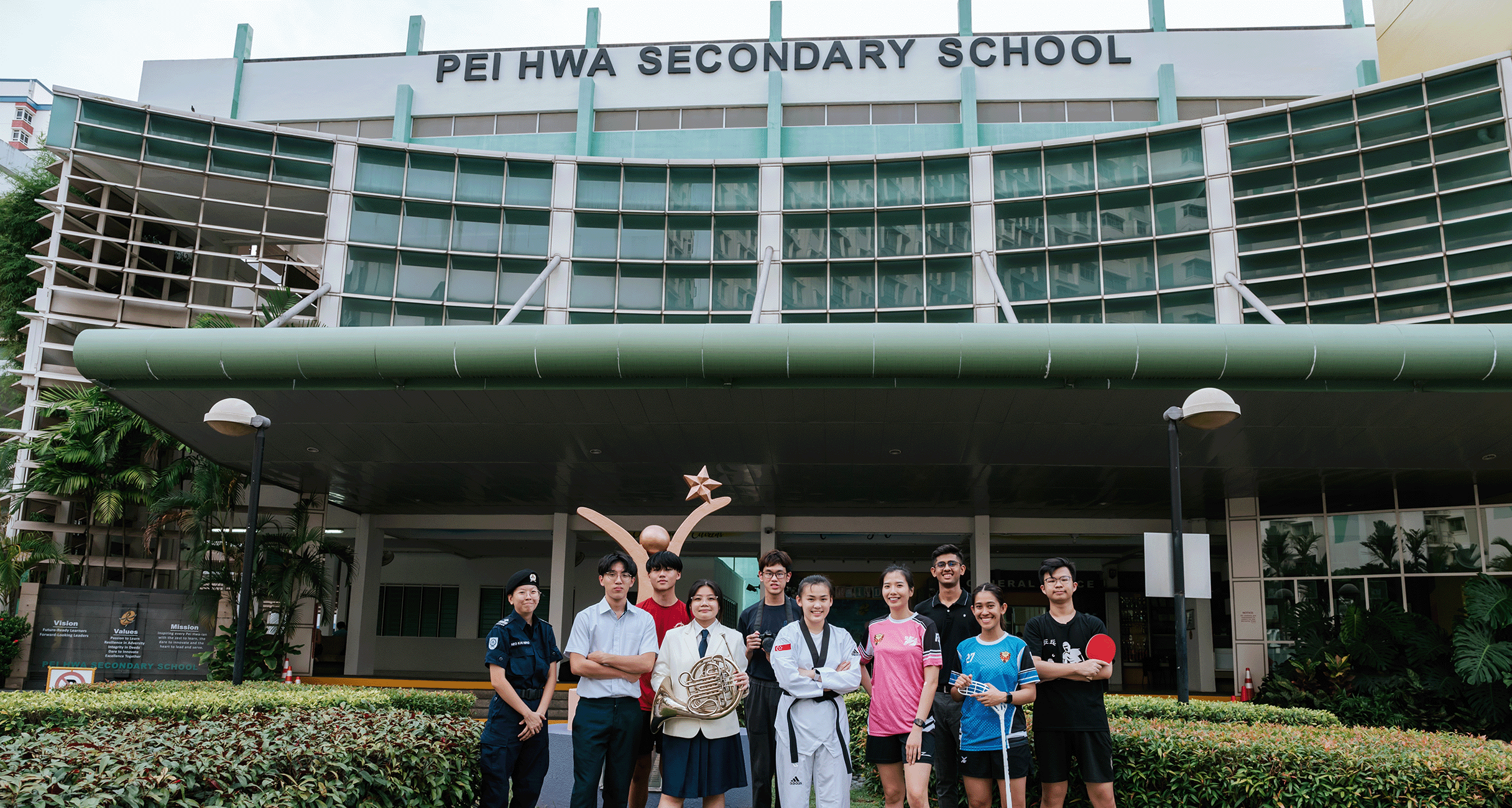 Students in various uniforms and sports attire pose in front of Pei Hwa Secondary School building.